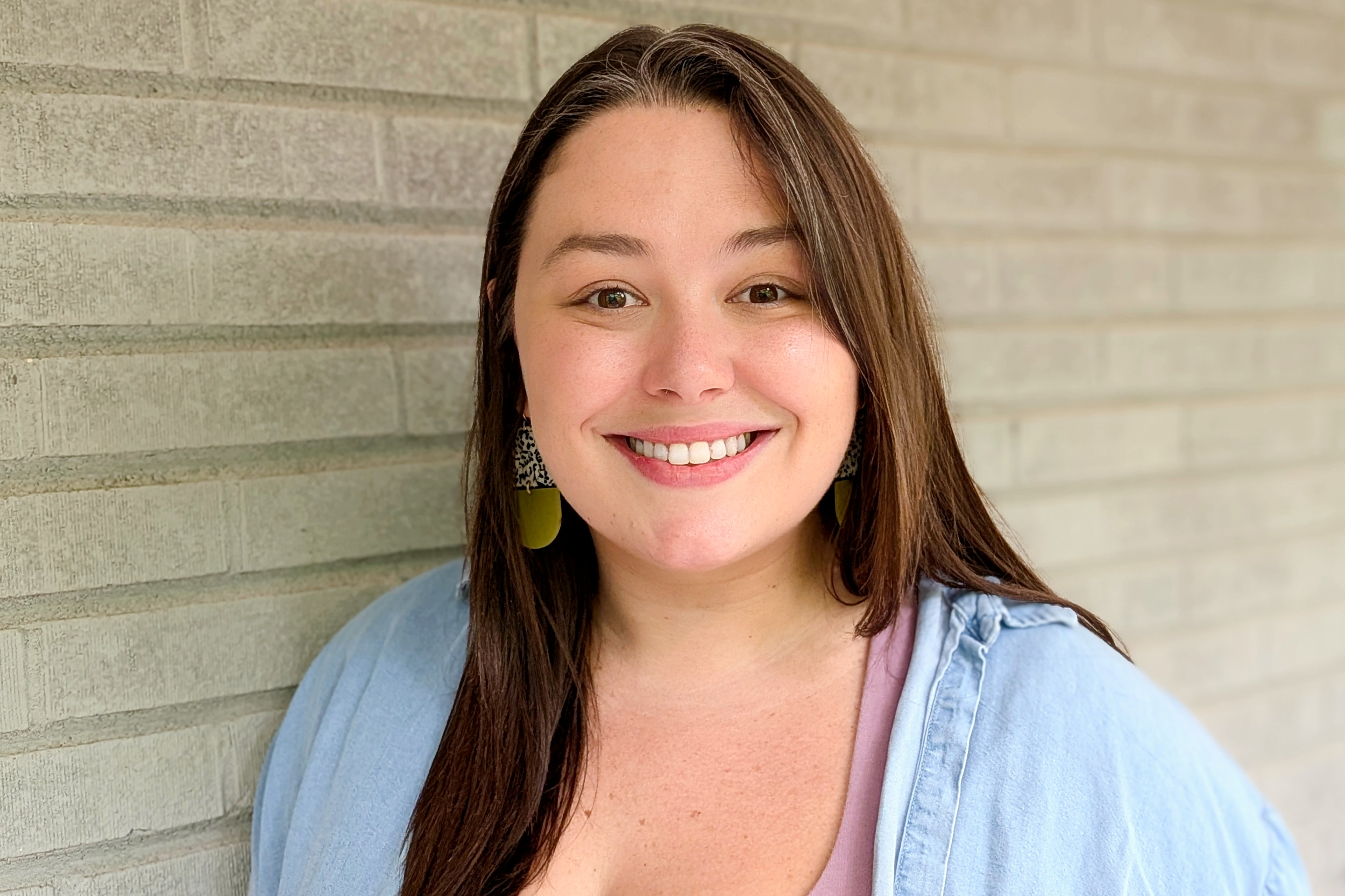 Smiling woman with long brown hair, wearing gold earrings and a denim jacket.