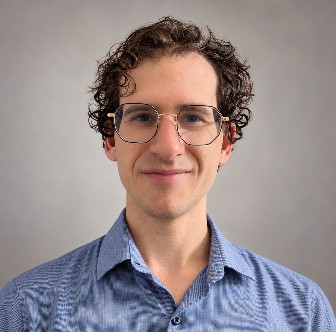A young male therapist with curly brown hair, wearing glasses and a blue dress shirt, smiling in front of a plain gray background.