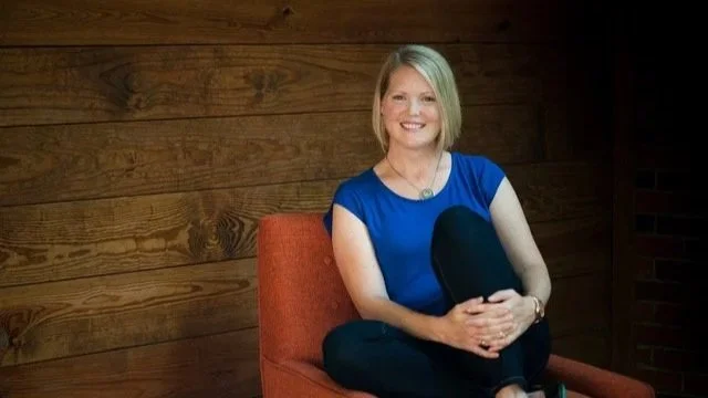 A female therapist with blonde hair smiling, sitting on an orange chair with a wooden wall background.