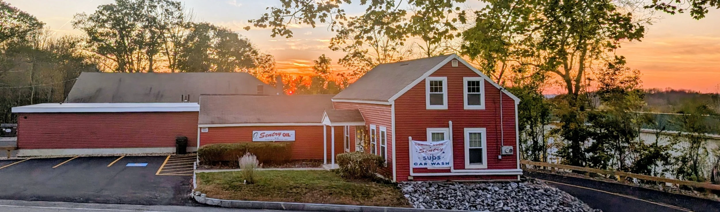 Red building with a sign for Sentry Oil and car wash, sunset in background, surrounded by trees.