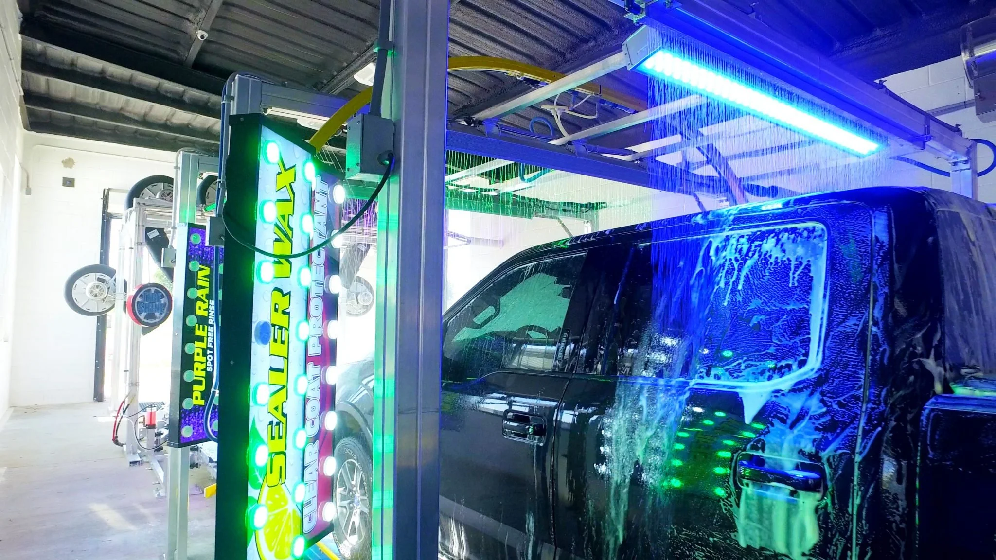 A black vehicle is being washed at a car wash station with multiple brushes and water jets, illuminated by blue and green neon lights.