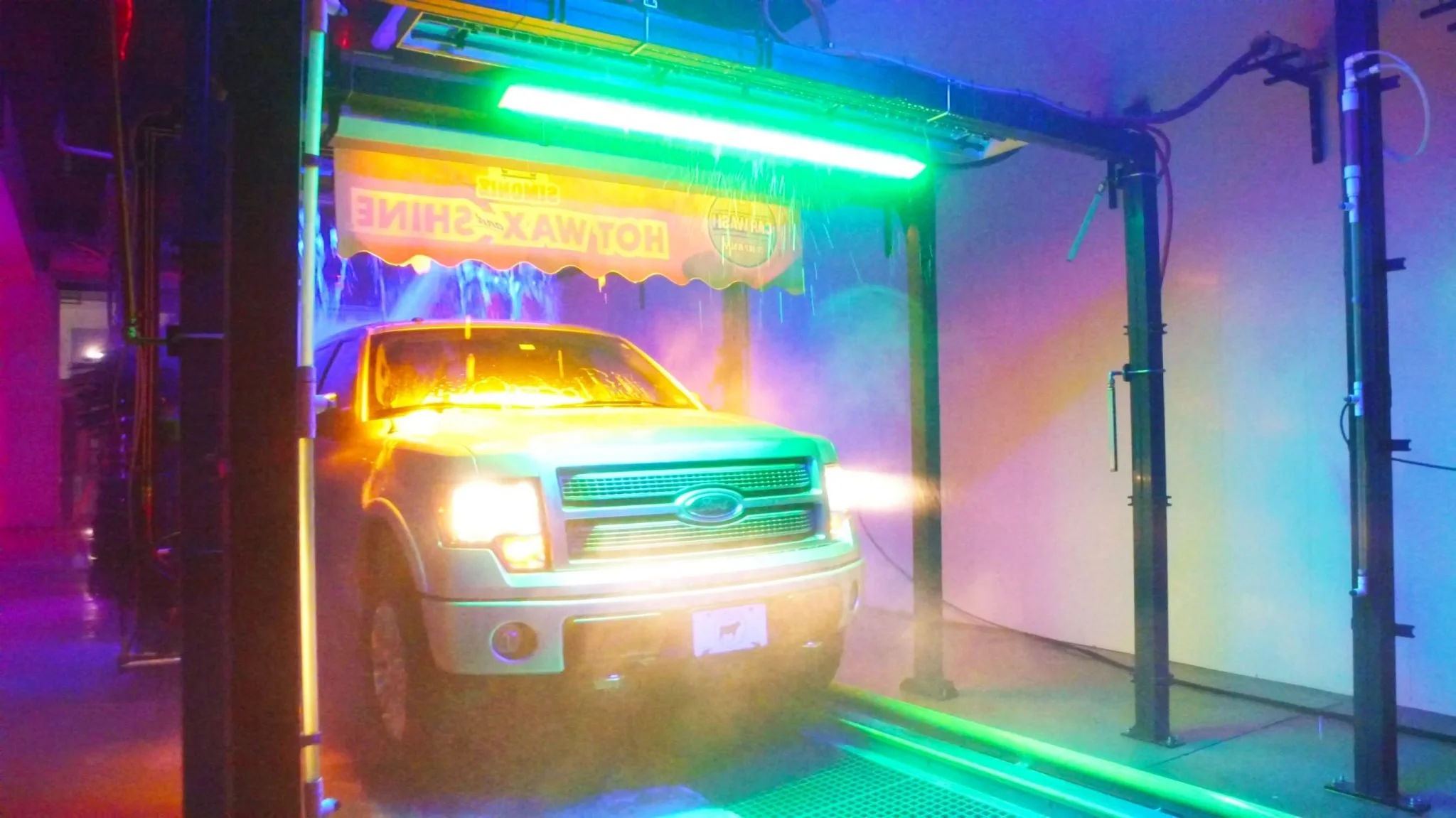 A silver Ford pickup truck in a car wash tunnel with colorful neon lights and soap suds in the air.