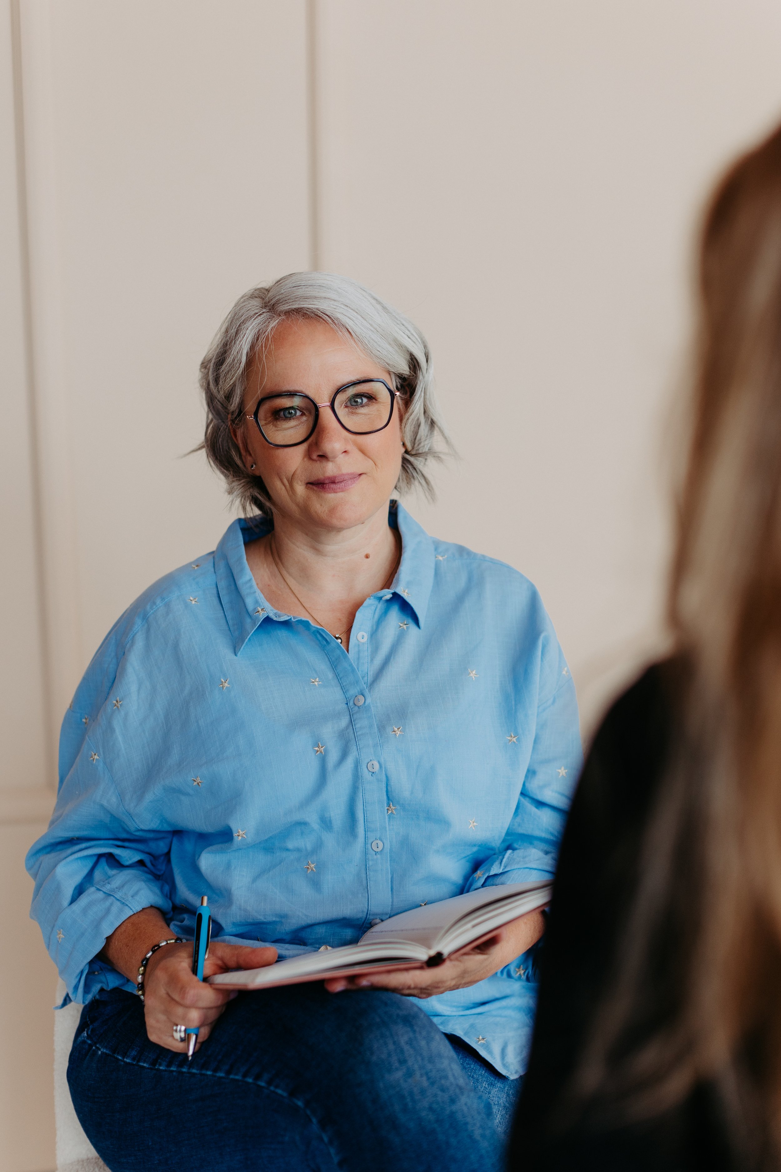 An woman with short gray hair, wearing glasses and a light blue shirt with star patterns, is sitting down, holding a notebook and pen, and engaging in conversation with a person who is partially visible in the foreground.