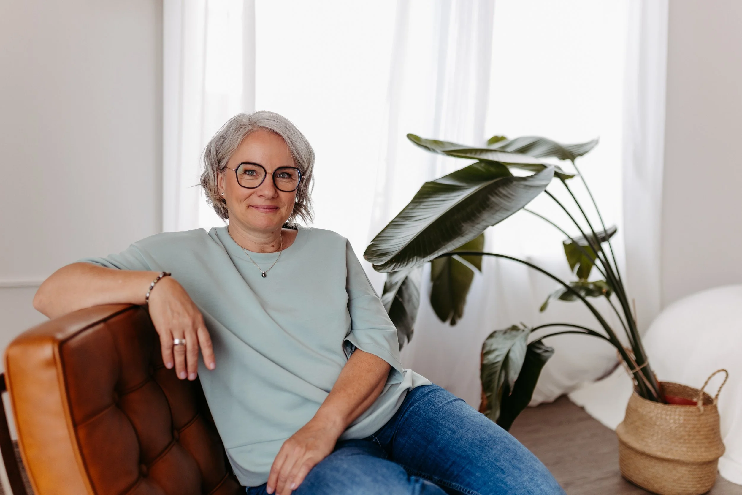 A smiling woman with gray hair and glasses sitting on a couch in a bright room, near a large potted plant, wearing a light blue top and jeans.