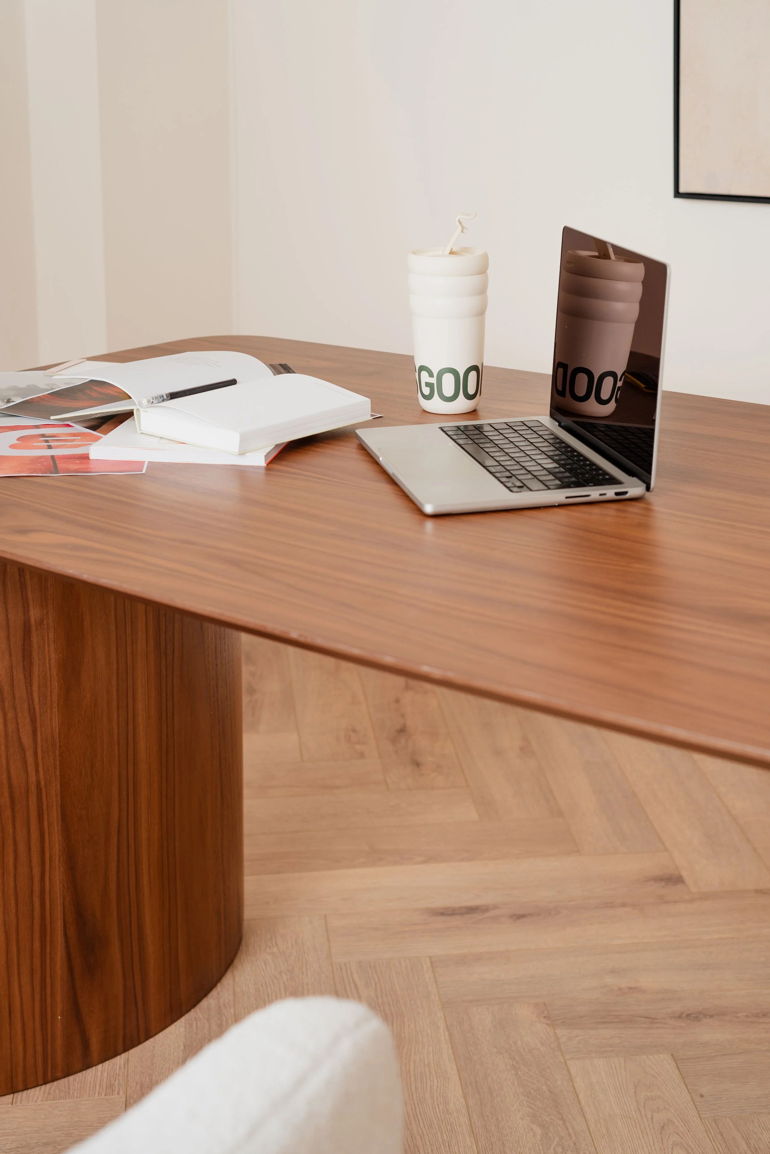 Wooden desk with an open notebook, magazines, a pen, a white travel mug with a straw and the word 'GOOD' on it, a closed laptop with a reflective screen, and a white upholstered chair in the foreground.