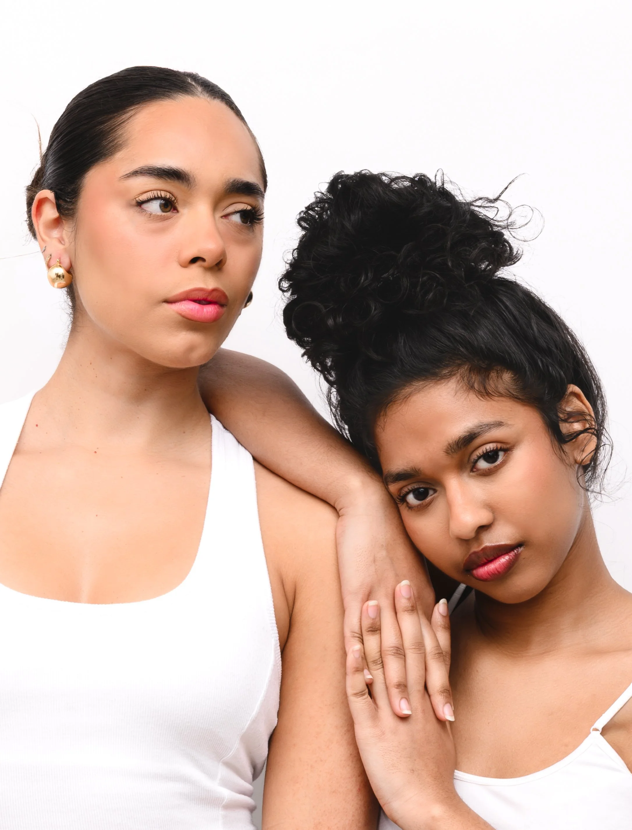 Two women with one woman resting her arm on the other's shoulder, both dressed in white. The woman on the left has dark hair pulled back and wears gold earrings, while the woman on the right has dark curly hair styled up and rests her hand on the other's arm.