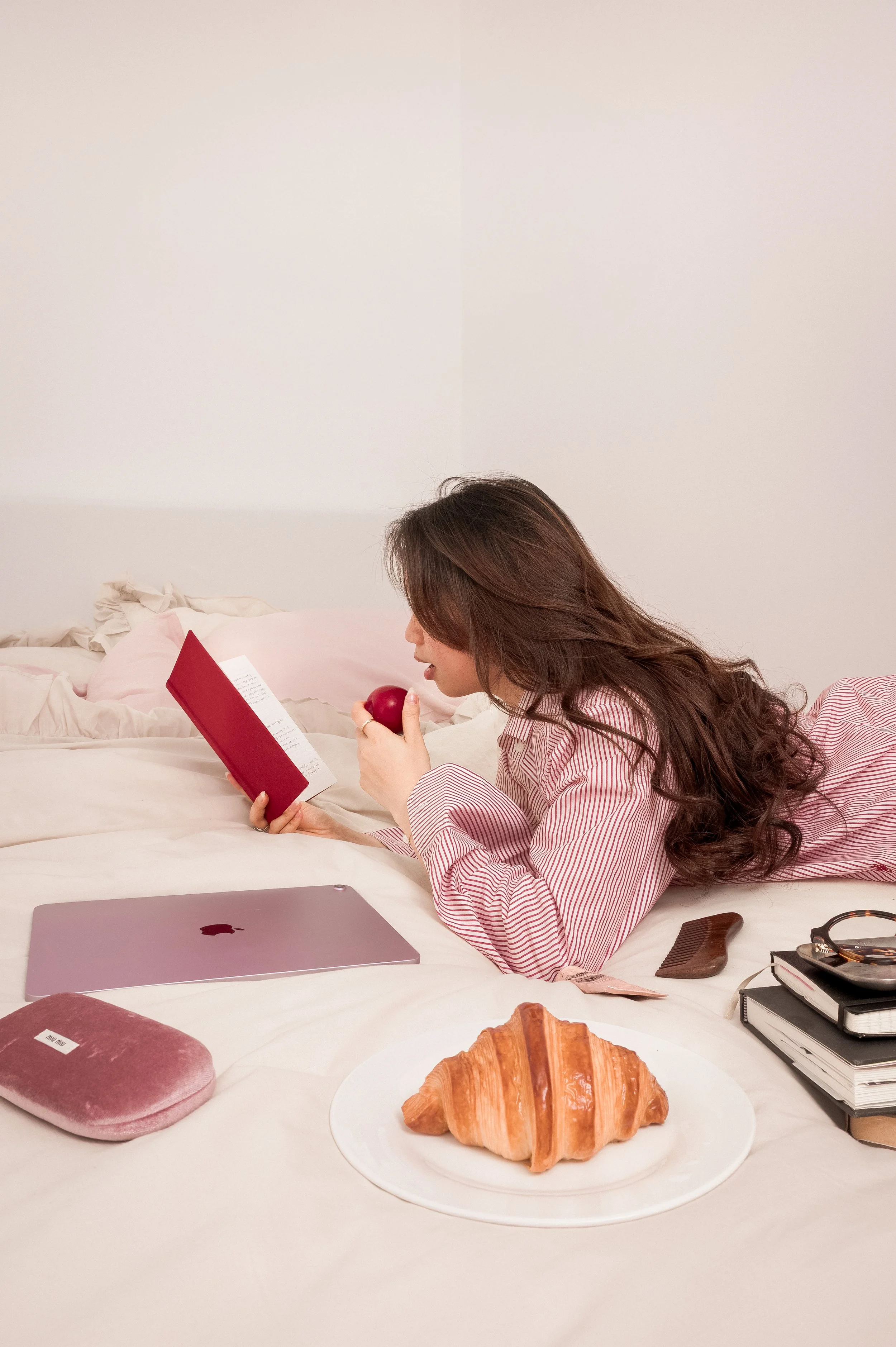 A woman lying on her stomach on a bed, reading a book while holding an apple. There is a plate with a croissant and various objects on the bed, including a pinked laptop, a pink velvet pouch, a hairbrush, notepad, and headphones.