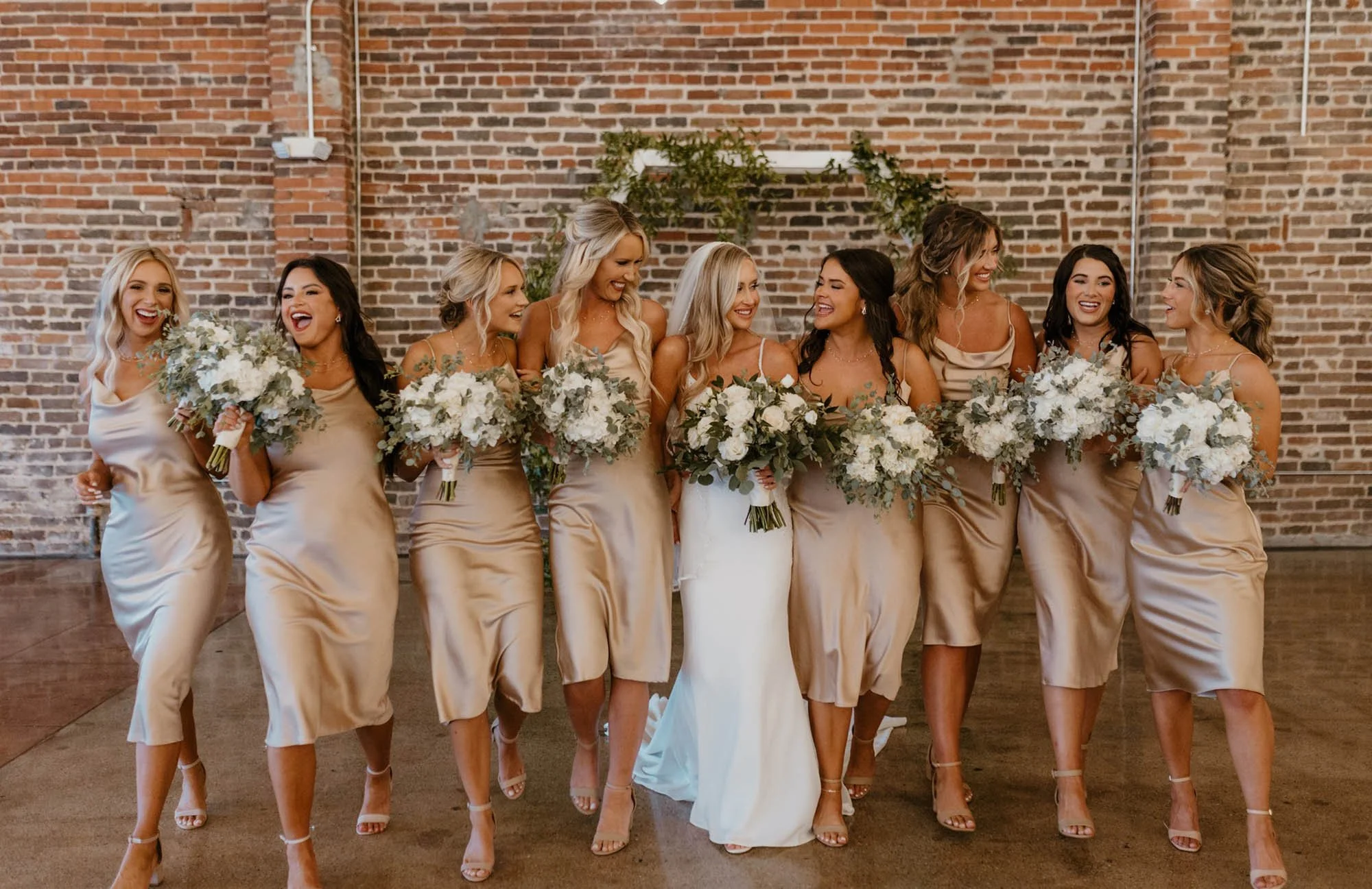 Bride and bridesmaids in beige dresses holding bouquets, walking in a line against a brick wall.