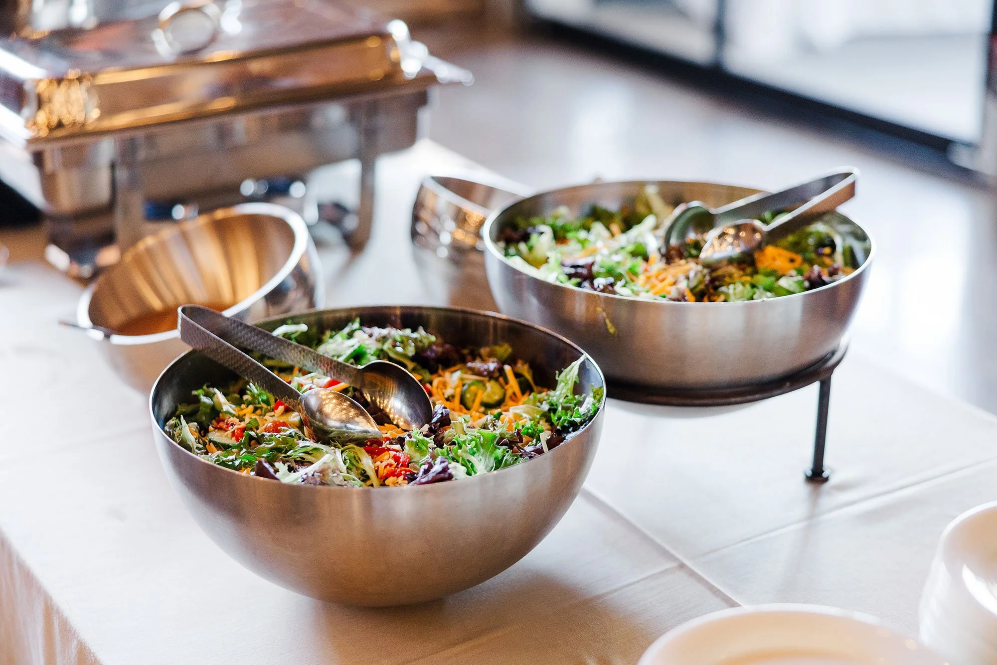 Two stainless steel bowls filled with salad on a buffet table, with salad tongs inside each bowl.
