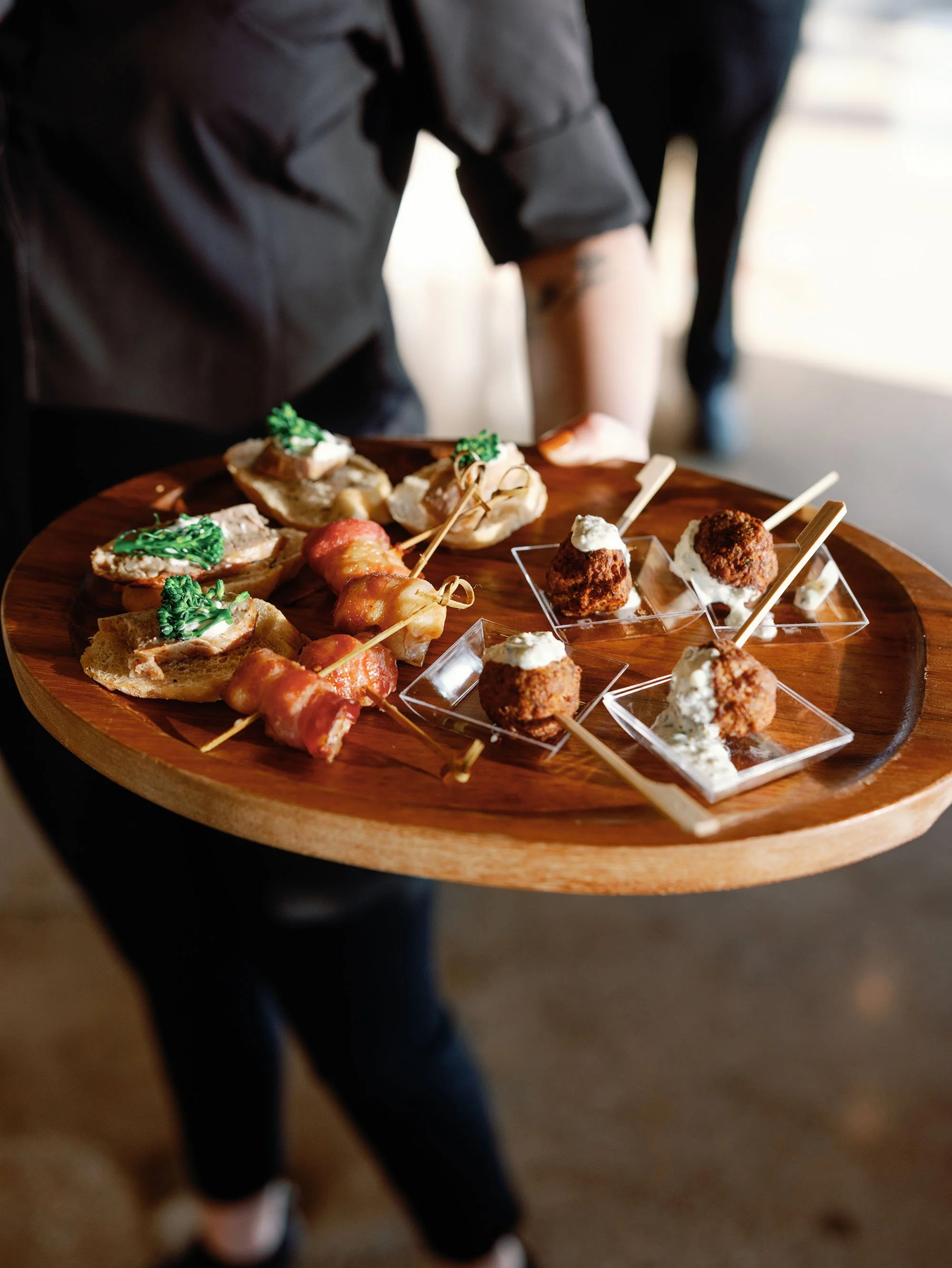 A server holding a round wooden tray with assorted appetizers, including small sandwiches with green garnish and skewered bacon-wrapped items, along with breaded meatballs topped with white sauce, on small clear dishes.
