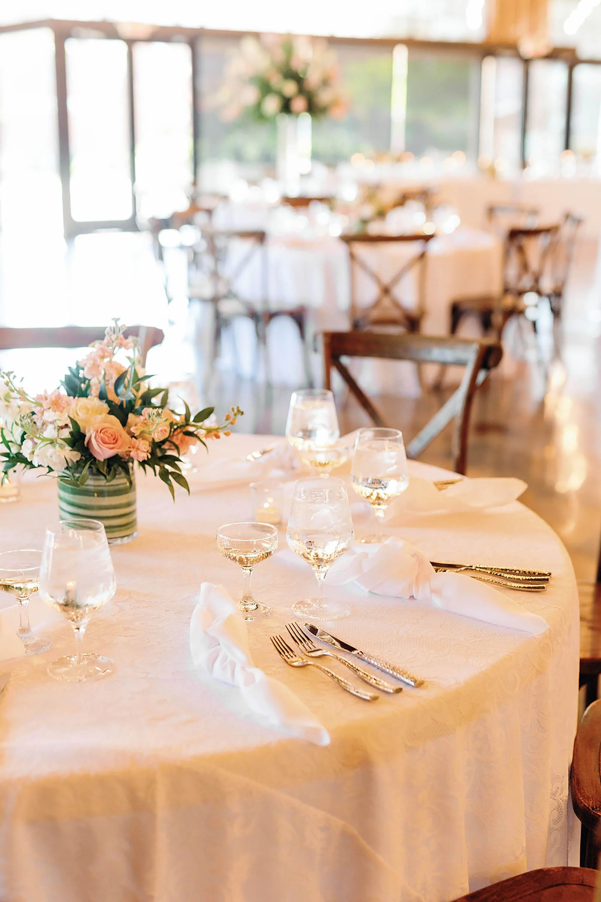 Elegant banquet table with white tablecloth, floral centerpiece, wine glasses, and neatly folded napkins, set in a bright decorated event space with multiple tables and chairs.
