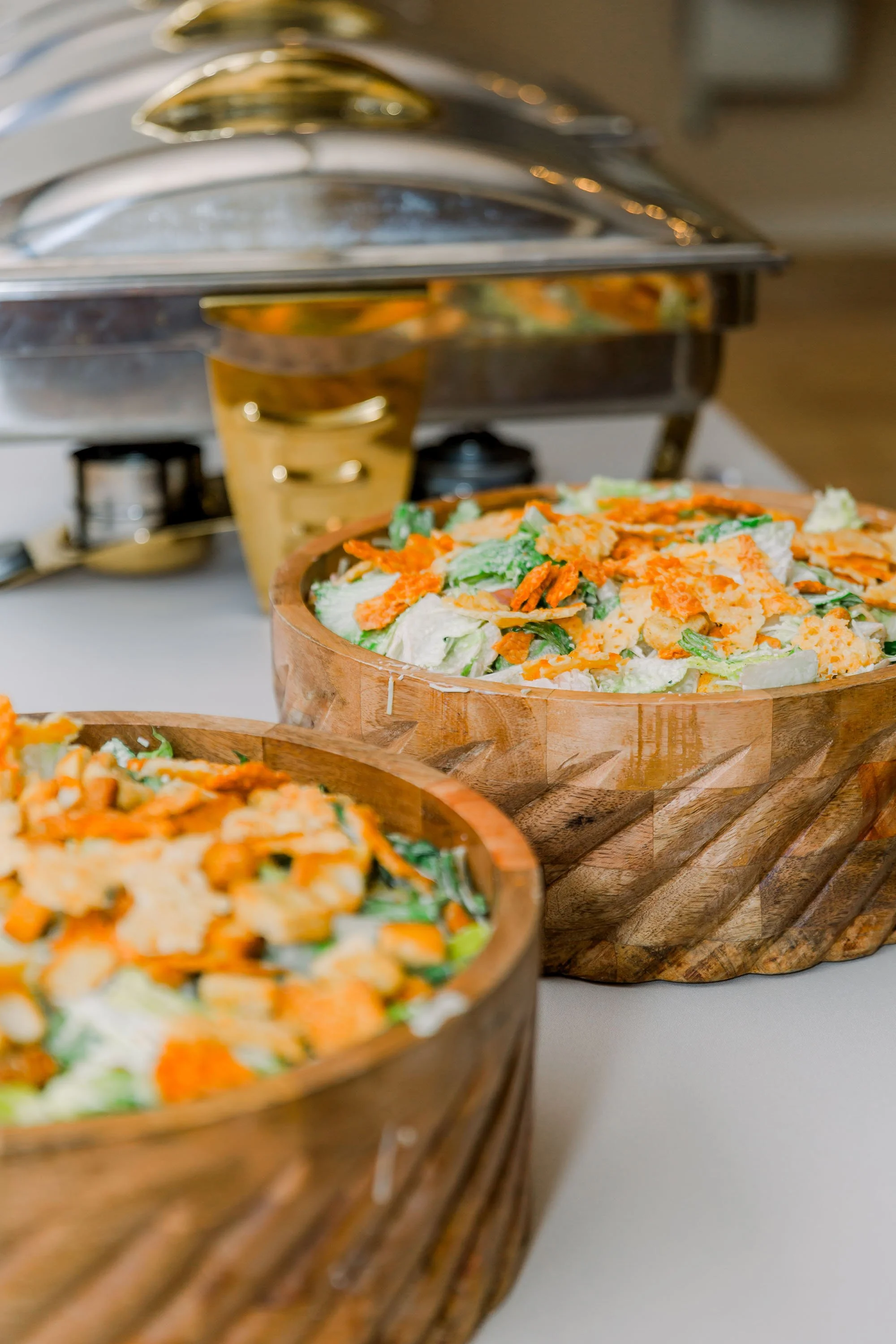 Two wooden bowls filled with salad on a table, with a chafing dish in the background.