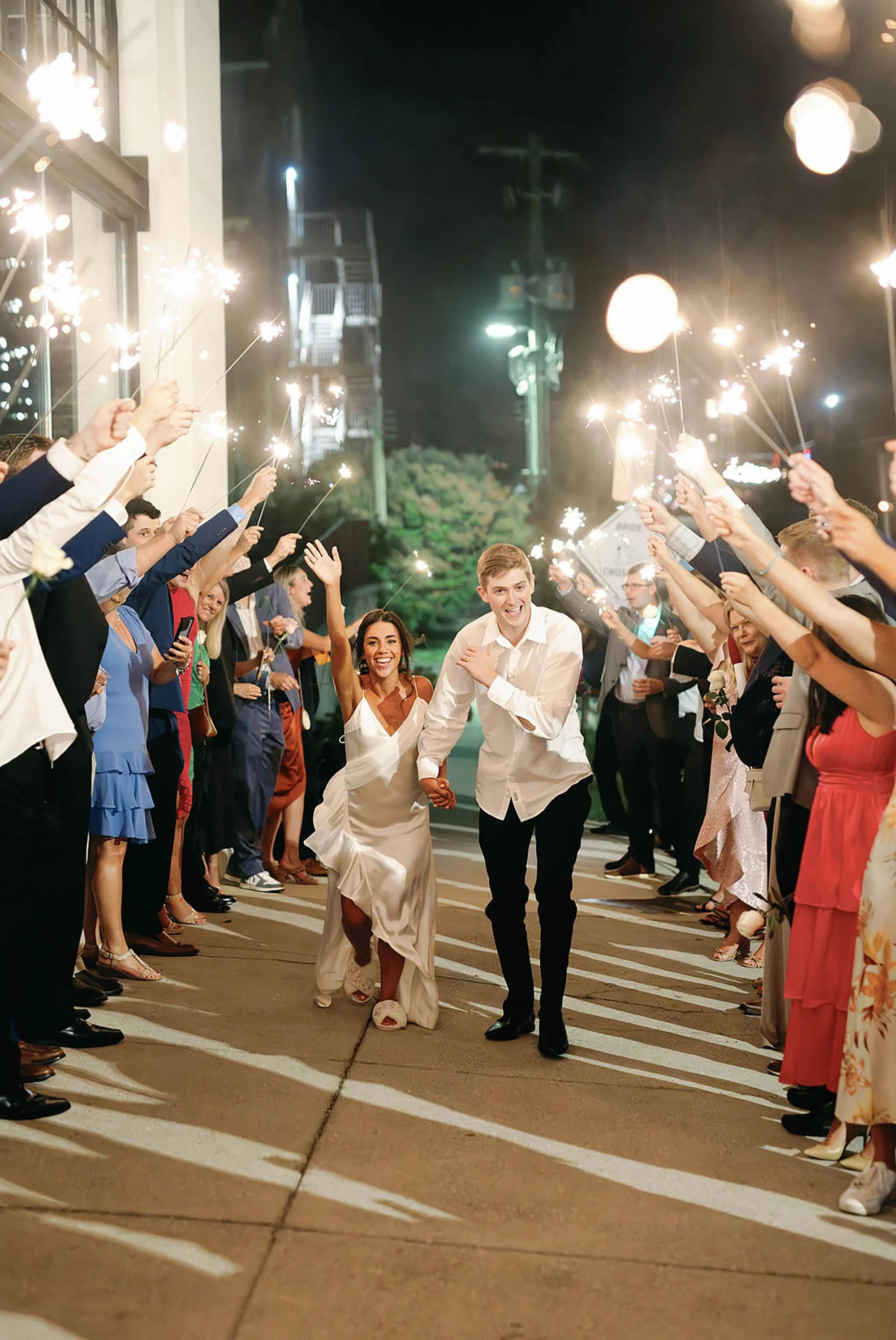 A joyful bride and groom run through a street lined with celebrating friends holding sparklers at night.