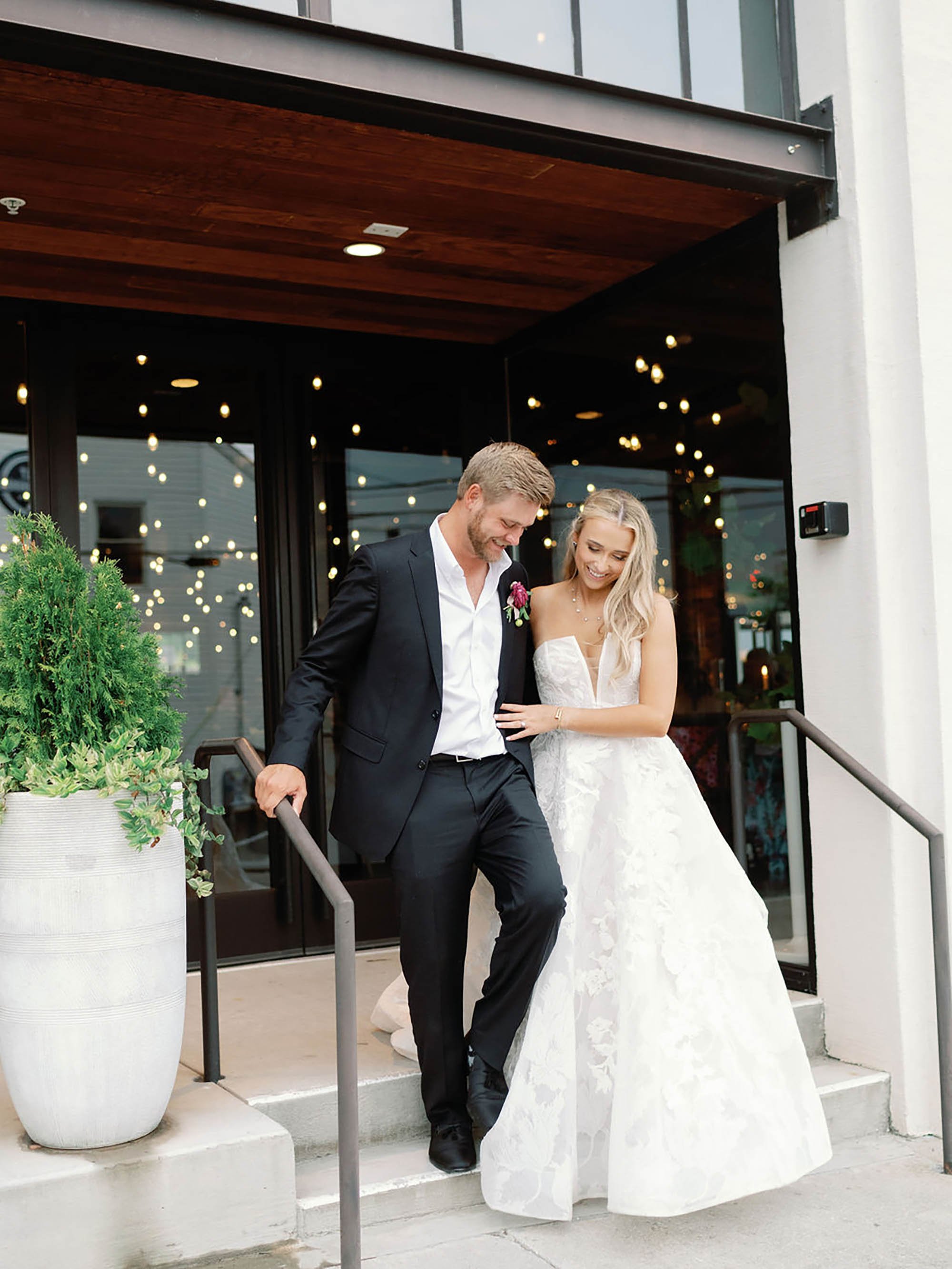 A newlywed couple walking down the steps outside a building, smiling and holding each other. The groom wears a black suit, and the bride wears a white wedding gown.