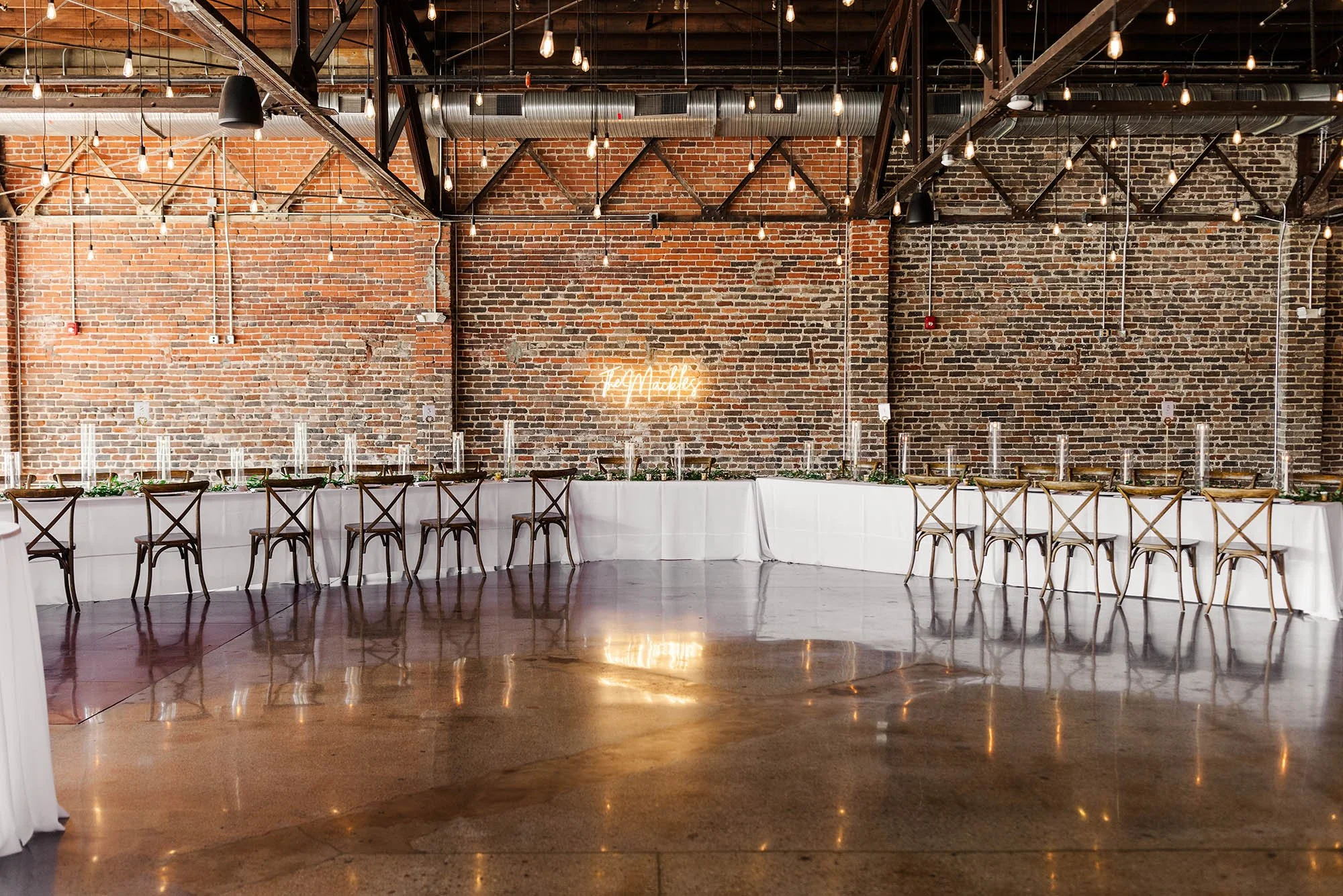 A long banquet table set for an event, with tall glass candle holders and greenery, against a rustic brick wall with a neon sign and string lights.