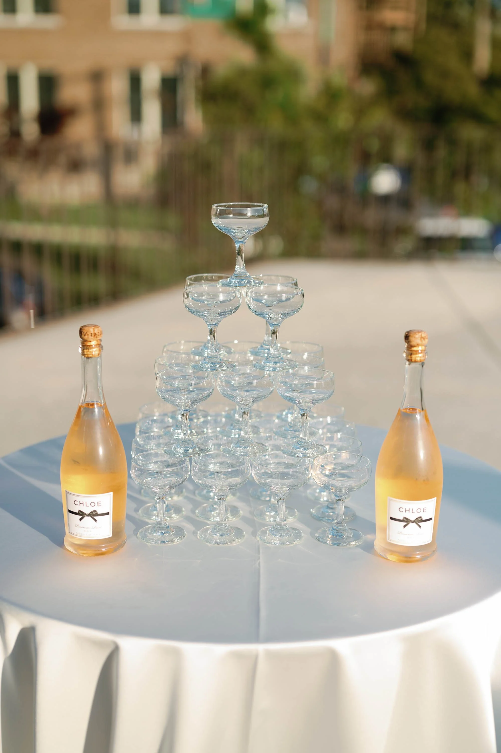 Champagne bottles and a pyramid of empty cocktail glasses on a round table at an outdoor event.