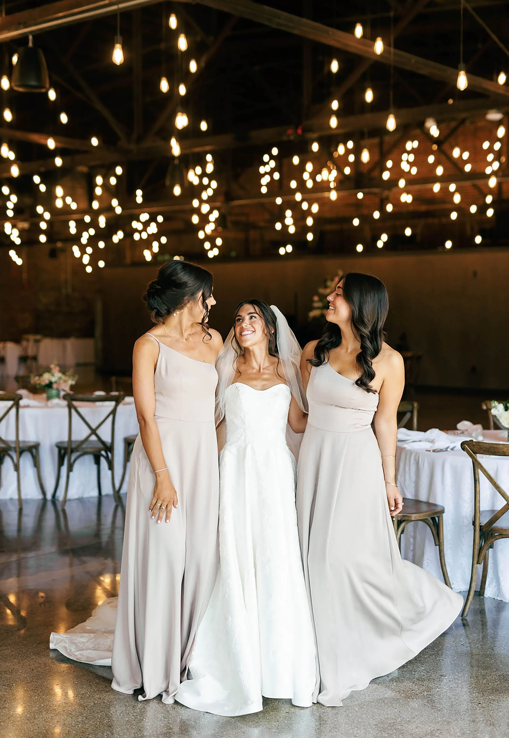 Three women at a wedding reception in a rustic indoor venue with hanging string lights. One is the bride in a white wedding gown with veil, and the other two are bridesmaids in beige dresses. They are smiling and looking at each other.