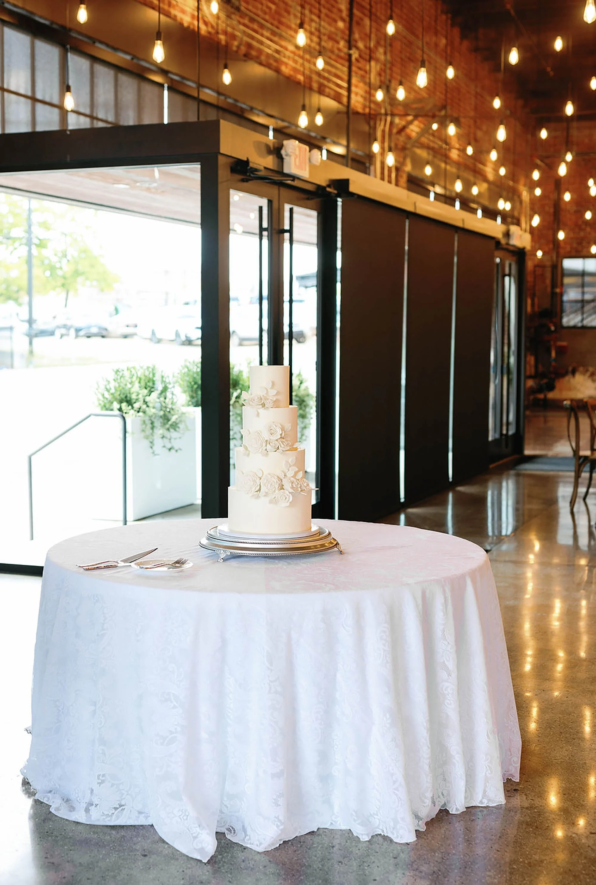 White wedding cake with floral decorations on a round table with a white tablecloth, inside a rustic venue with warm lighting and large windows.