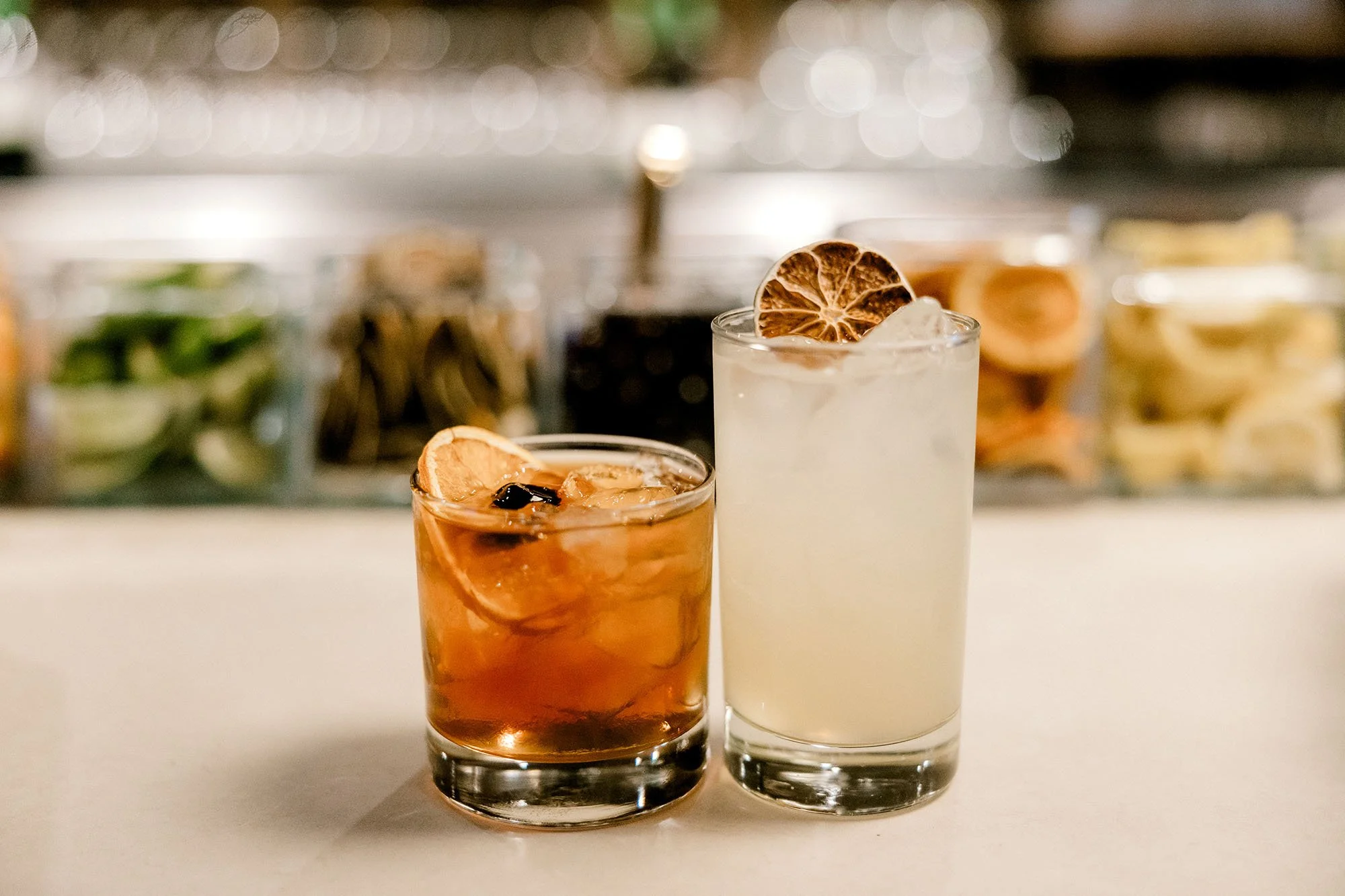 Two cocktails with dried citrus garnishes on a white surface, with blurred containers of fruit and herbs in the background.