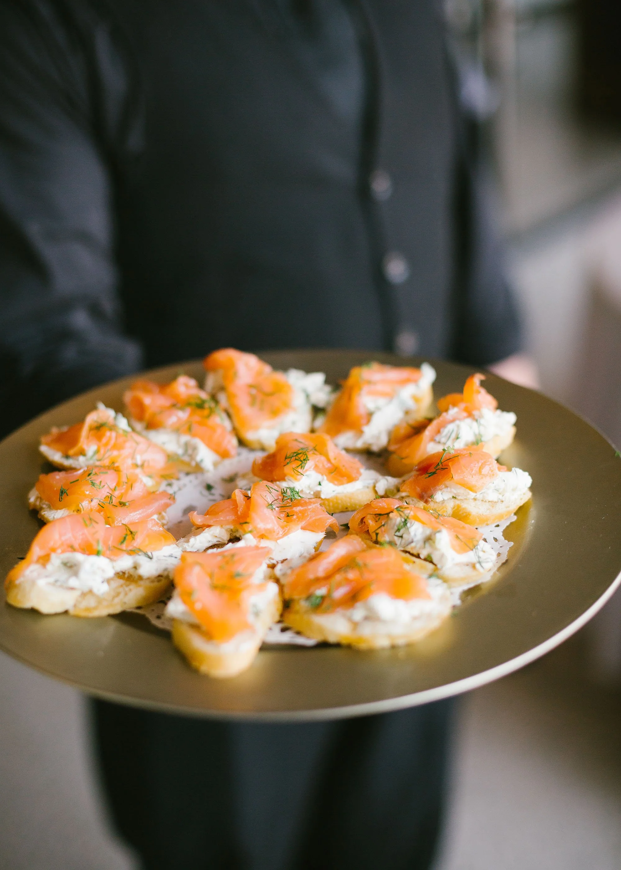Person holding a gold plate with 12 pieces of smoked salmon on toast topped with herbs.