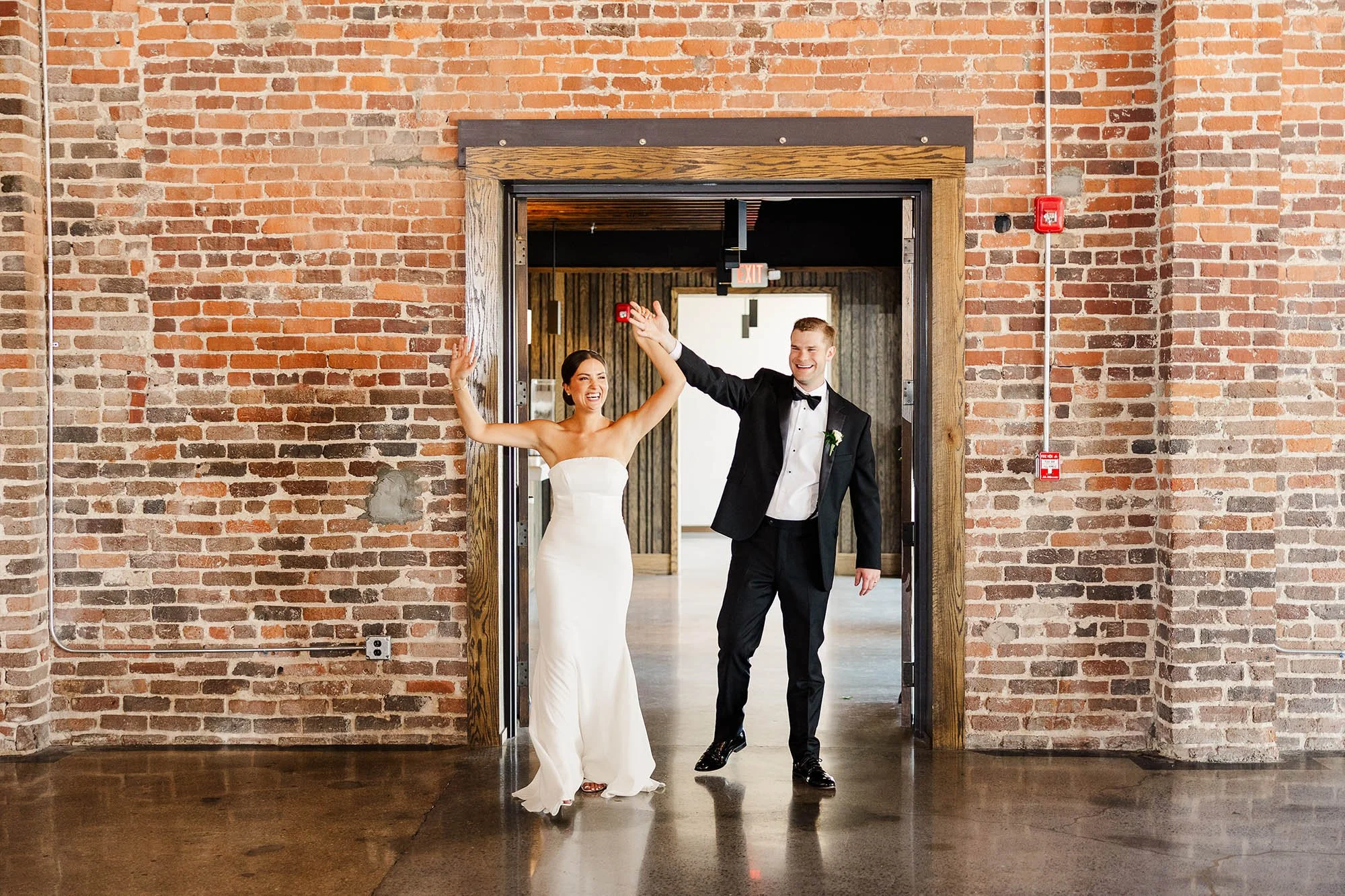 A bride and groom walking through a doorway, smiling and celebrating at their wedding reception in a rustic venue with exposed brick walls.