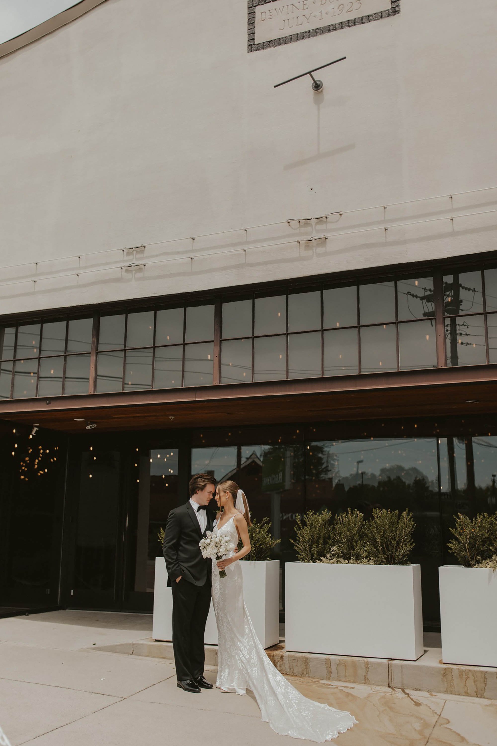 Bride and groom dressed in wedding attire standing closely outside a modern building, with the groom in a black tuxedo and the bride in a white lace wedding gown holding a bouquet of flowers.