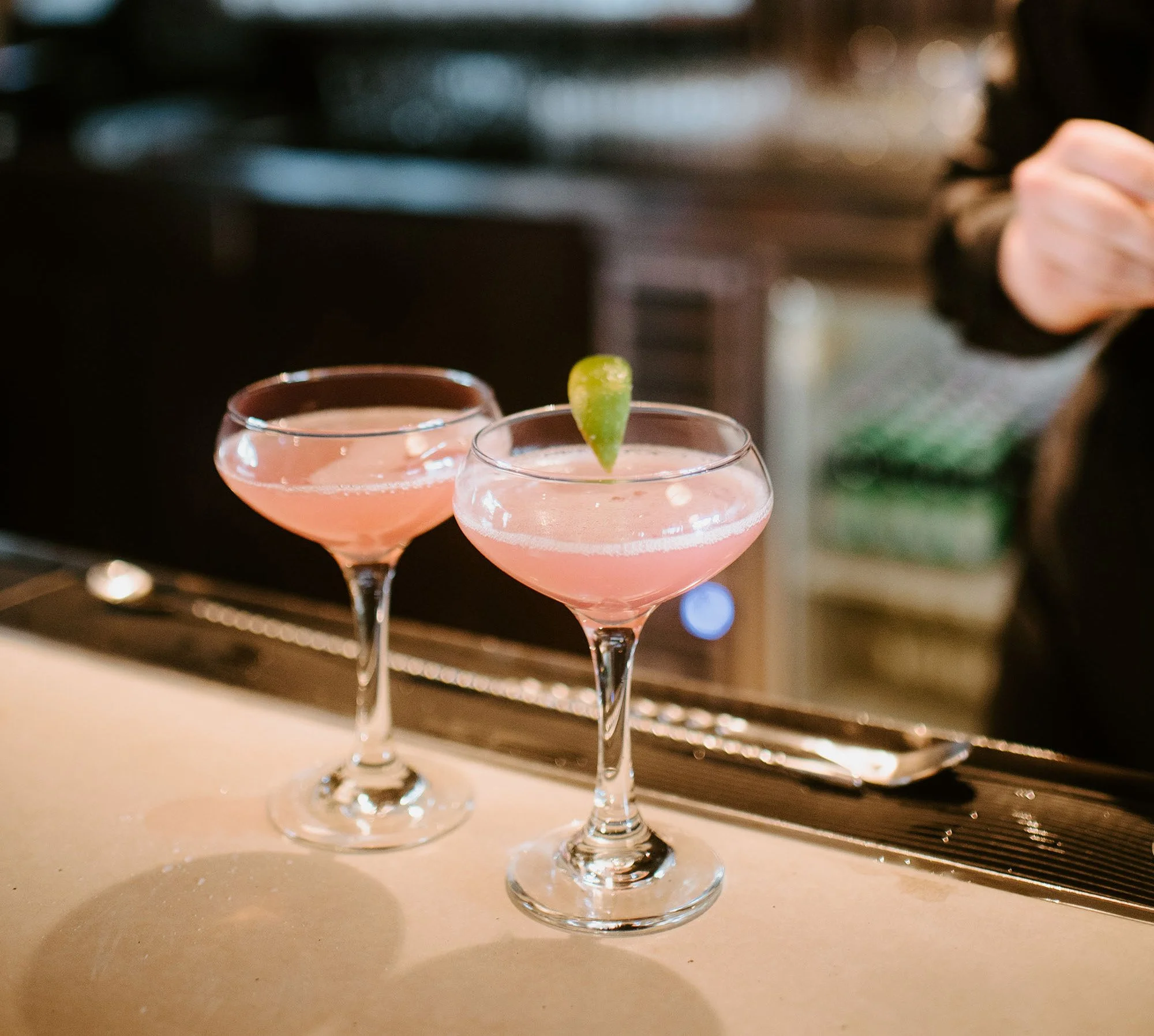 Two pink cocktails in coupe glasses, one garnished with a lime wedge, on a bar counter.