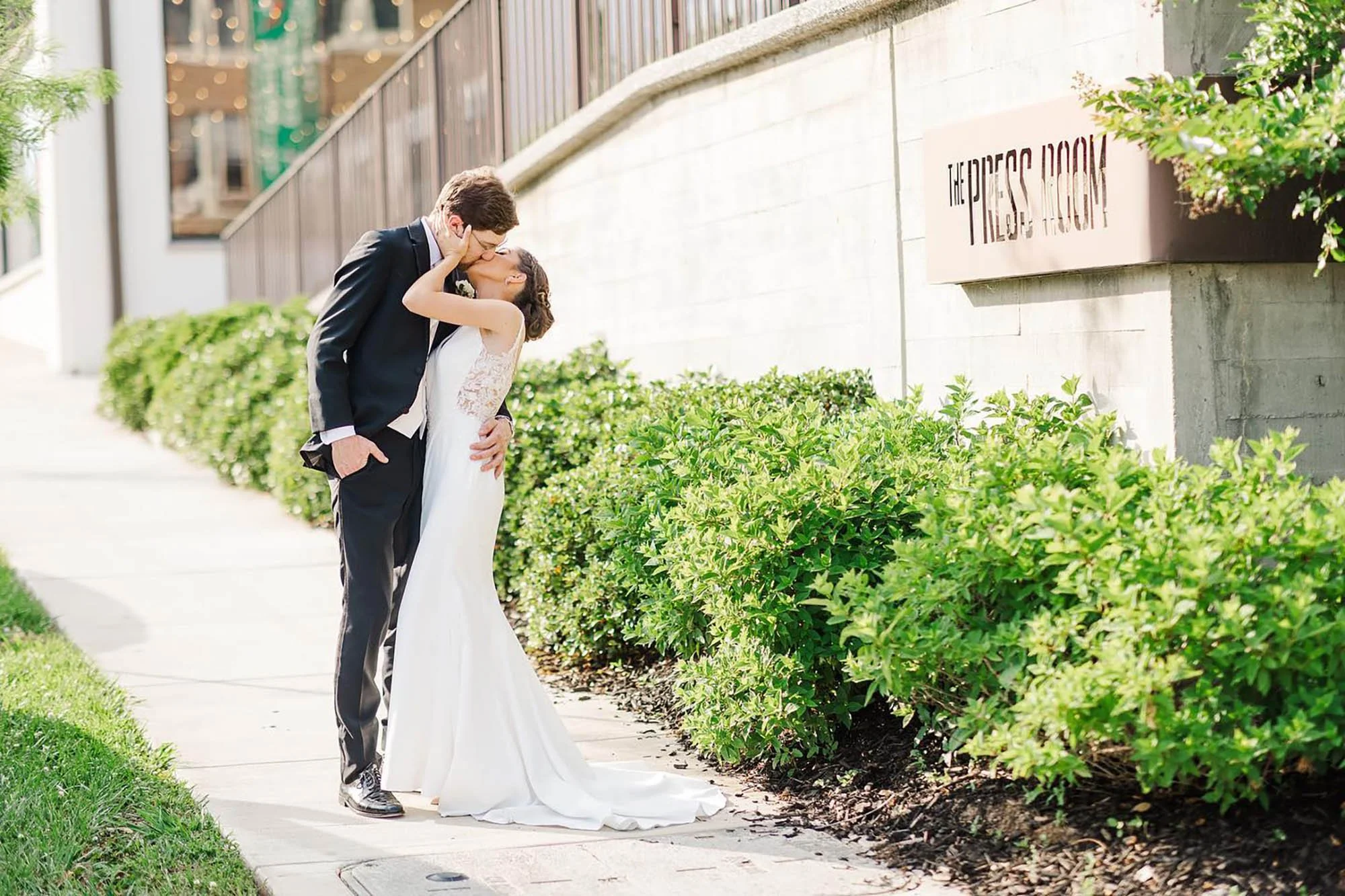 A bride and groom sharing a kiss outdoors on a sidewalk, with greenery and a wall with a sign that reads 'The Dress Room' in the background.