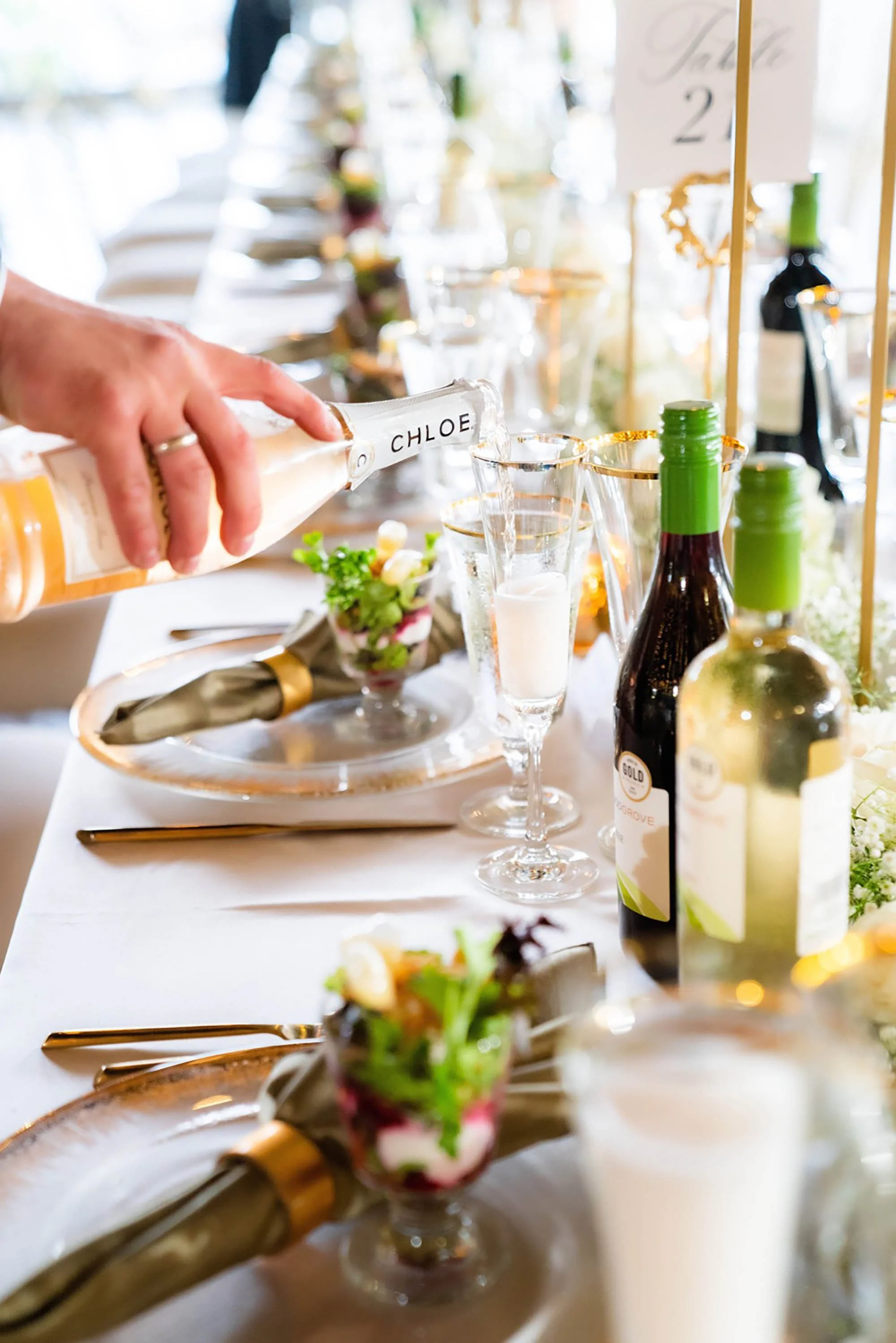A close-up of a dining table set for a formal event, with a person's hand pouring a drink into a glass, surrounded by floral arrangements, champagne glasses, wine bottles, gold-edged plates, silverware, and napkins.