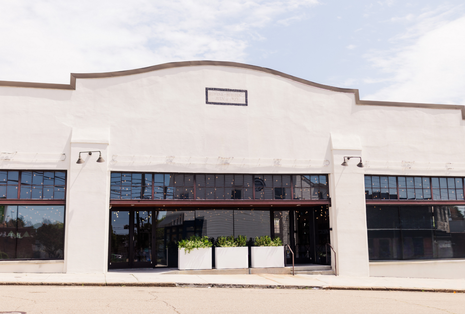 Front view of a white building with large glass windows, planters with greenery, and string lights inside, under a partly cloudy sky.