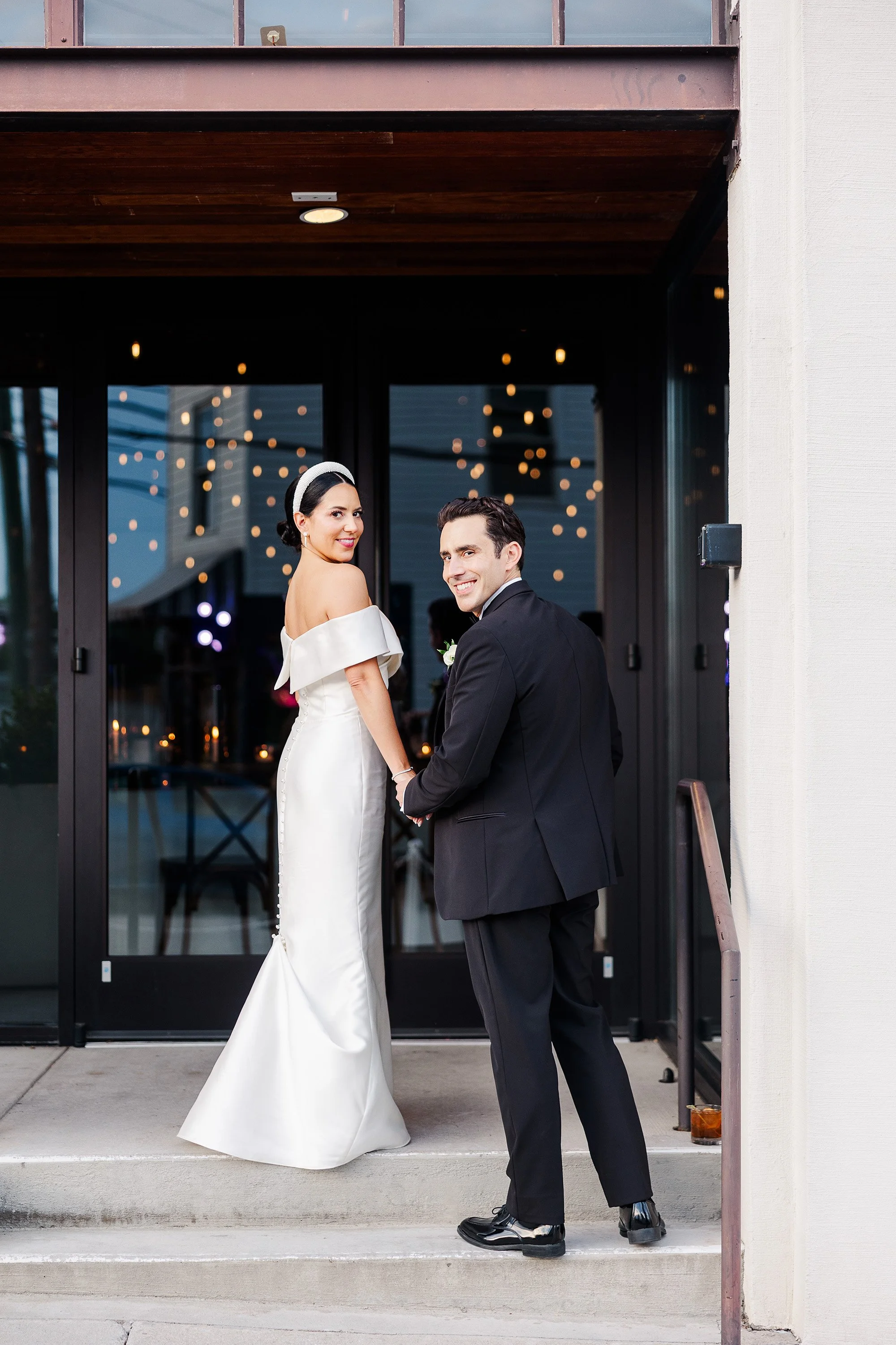 A bride and groom holding hands and smiling outside a building with glass doors and string lights reflected in the glass. The bride is wearing a white off-shoulder wedding gown with buttons down the back and a headband, and the groom is dressed in a 