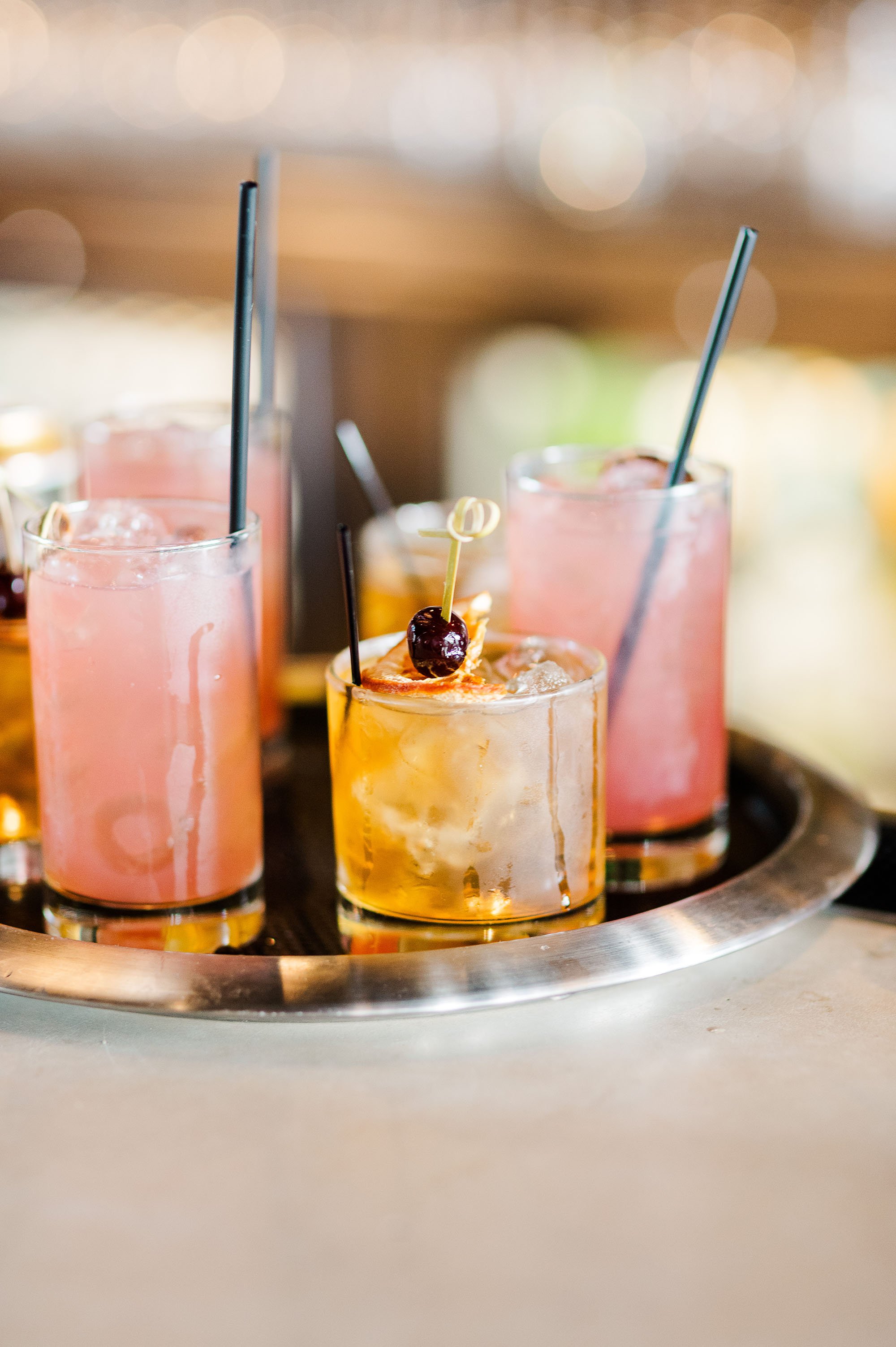 Three glasses of pink and yellow cocktails with ice and garnishes, on a silver tray.
