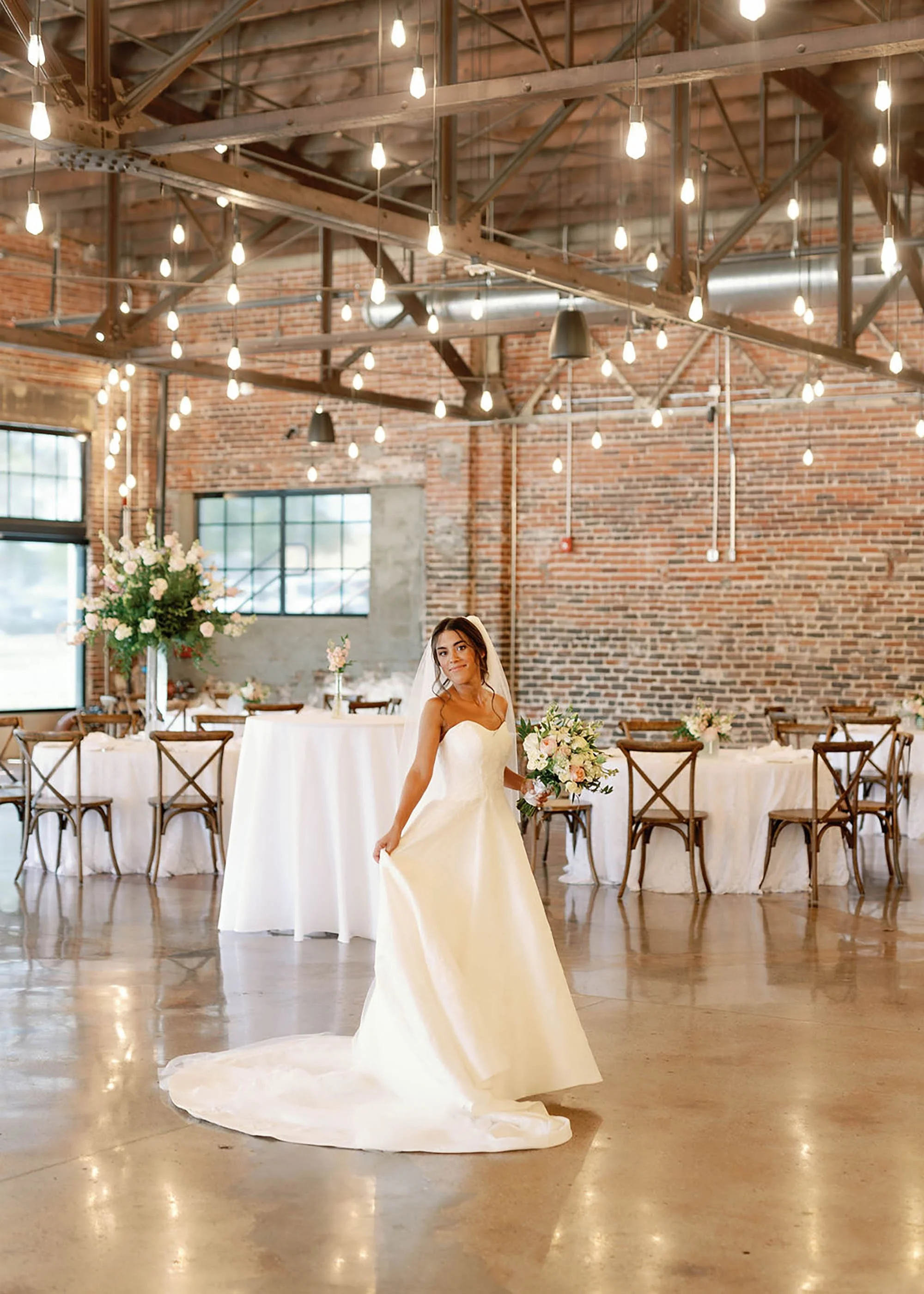 A bride in a satin wedding gown holding a bouquet of pink and white flowers, standing in a rustic industrial wedding venue with exposed brick walls, string lights, and decorated tables.