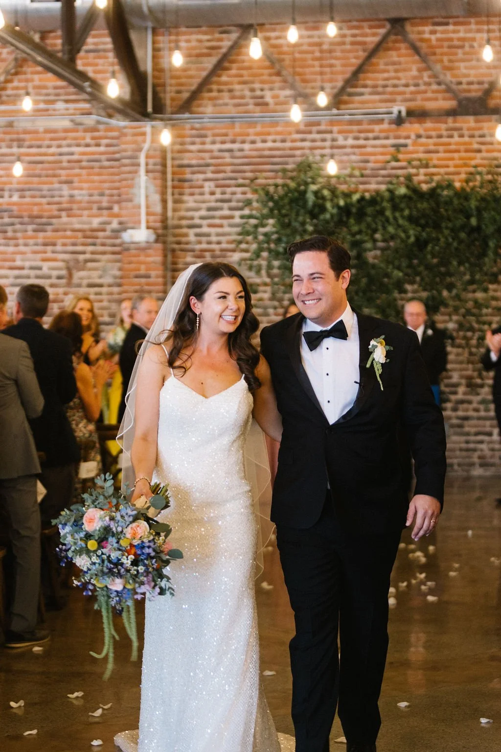 A newlywed couple, a woman in a white wedding gown holding a bouquet of flowers and a man in a black tuxedo, happily walking together at their wedding reception in a decorated venue with hanging lights and brick wall background.