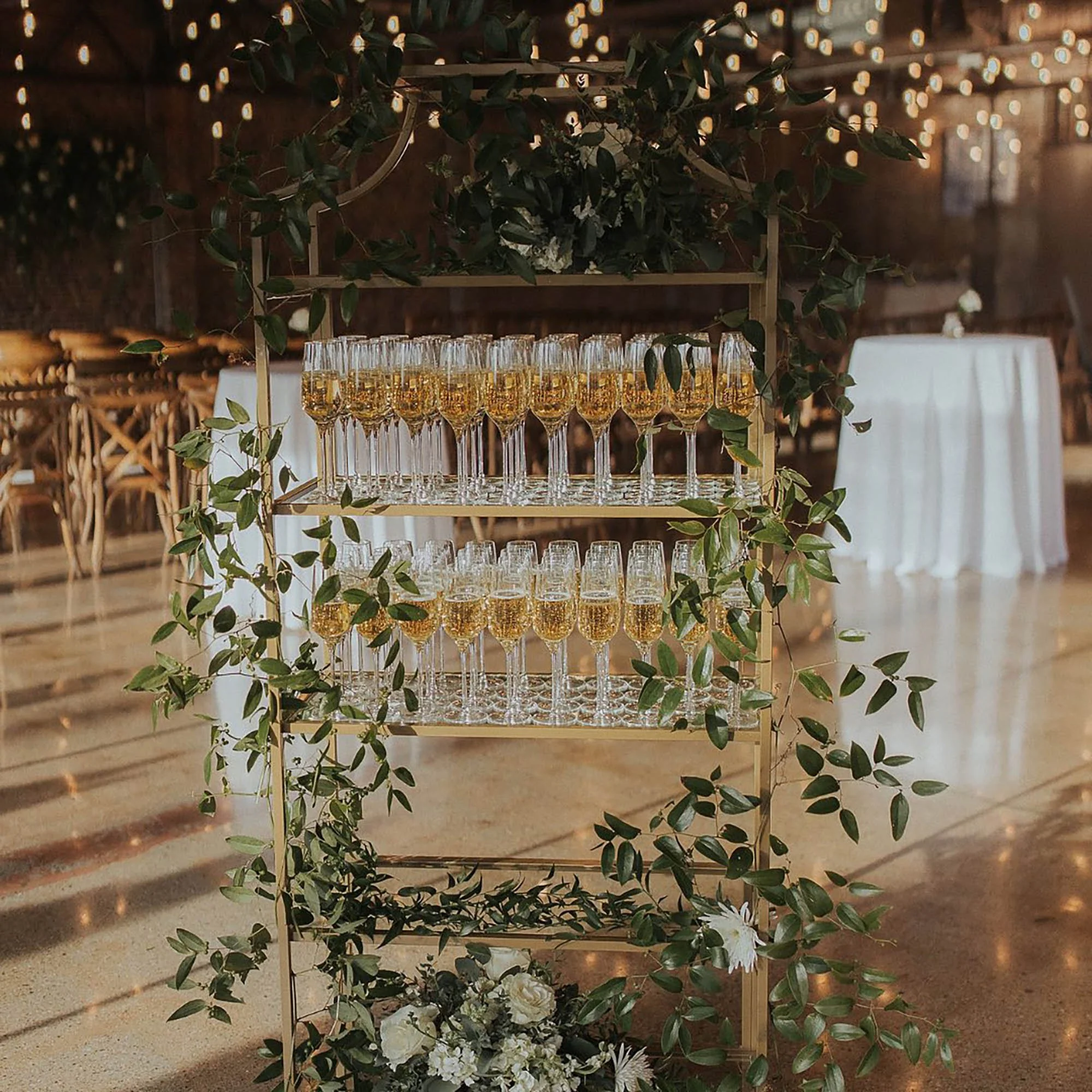 a decorative shelf with champagne glasses filled with a golden beverage, adorned with green leaves and white flowers, in a rustic indoor venue decorated with string lights