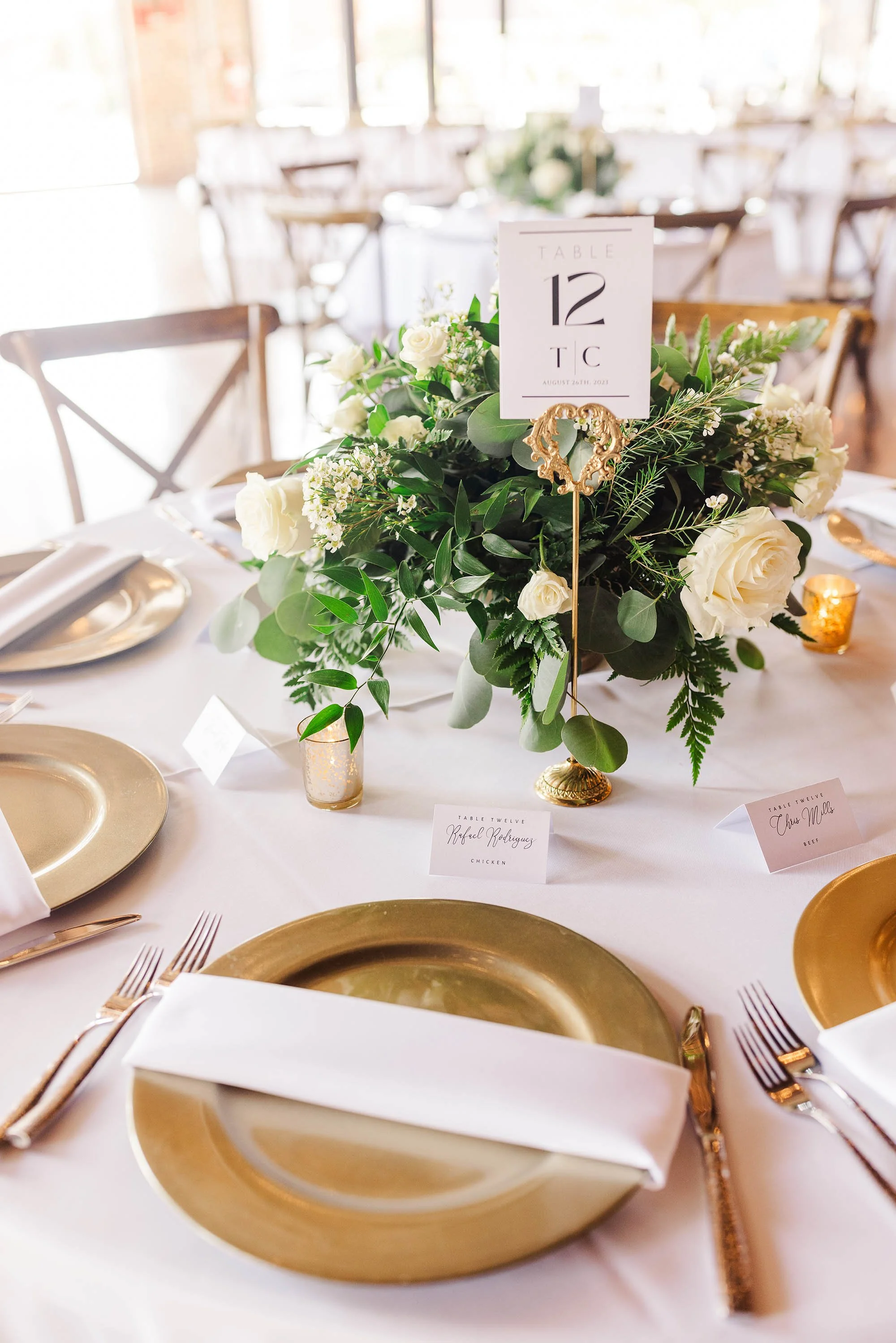 A wedding or formal event table setting with gold plates, gold utensils, white napkins, a floral centerpiece with white roses and greenery, table number card, and small votive candles.