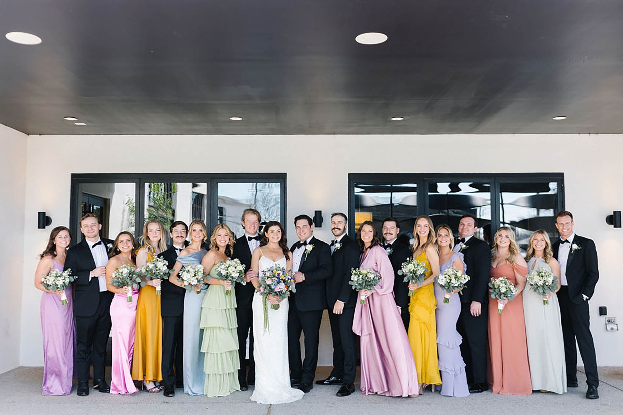 A group of wedding couples and bridesmaids standing together in front of a modern building, dressed in formal attire and holding bouquets of flowers.