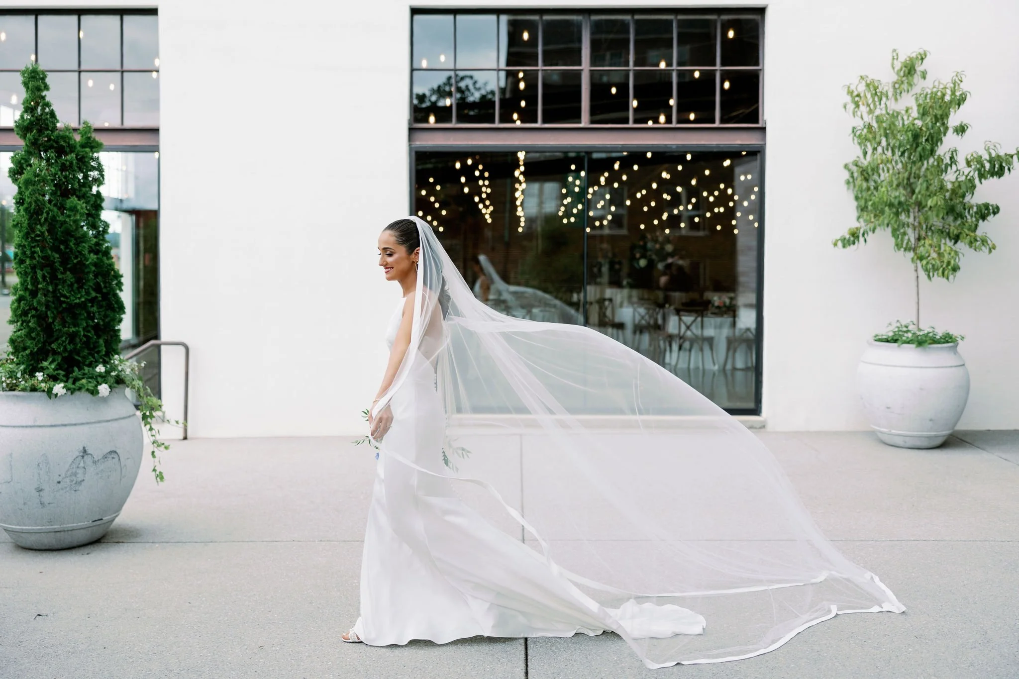 Bride in a white wedding gown and veil, smiling outside a modern building with large glass windows and string lights indoors.