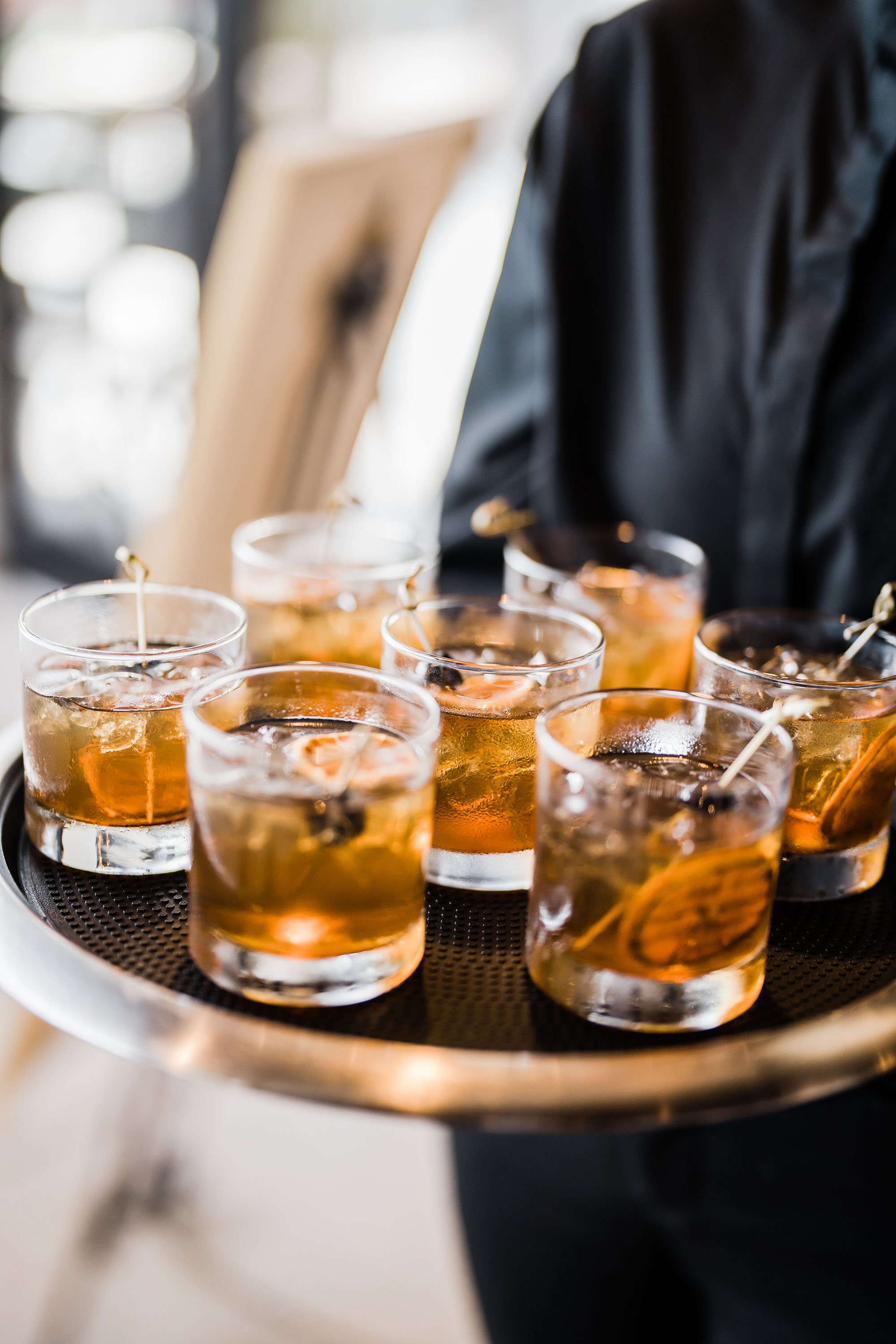 A server holding a tray of six glasses filled with an amber-colored drink, each garnished with a thin slice of orange.