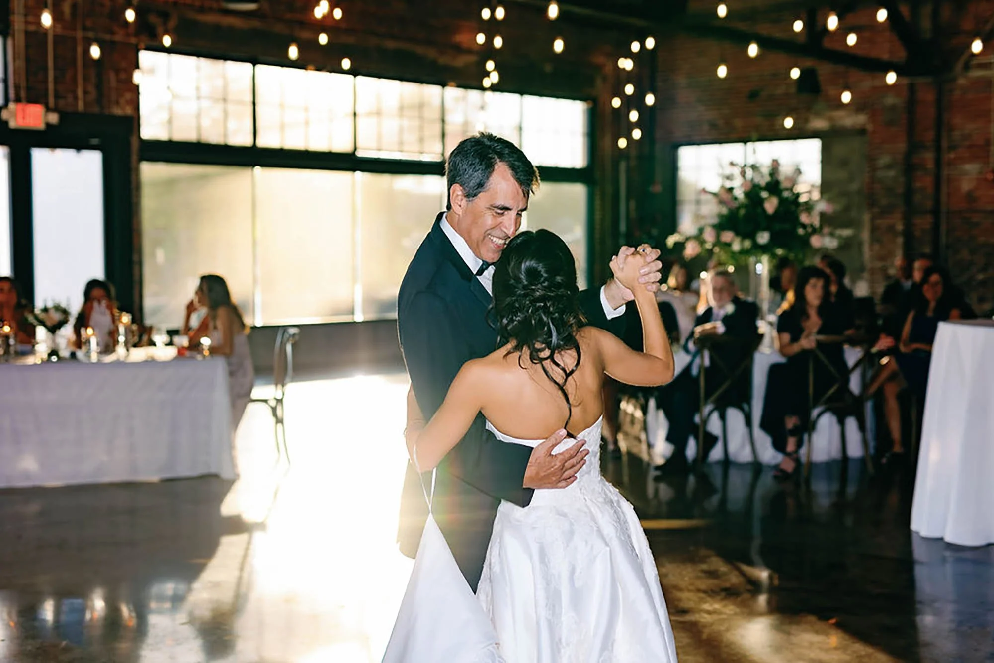 A man and woman dancing at a wedding reception, with guests seated at tables in the background and string lights hanging from the ceiling.