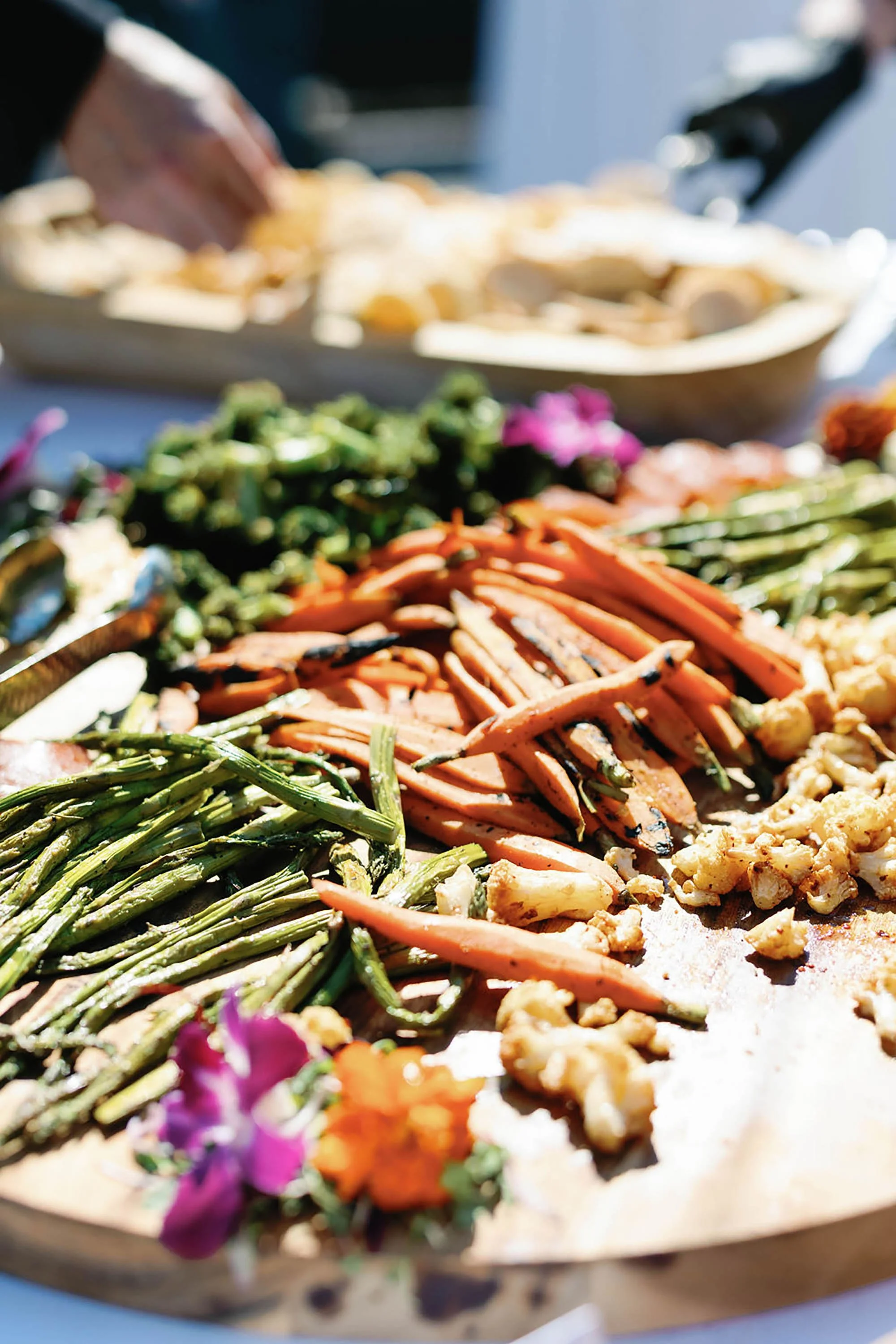 Assorted grilled vegetables including asparagus, carrots, cauliflower, and bell peppers on a wooden serving tray at a buffet.