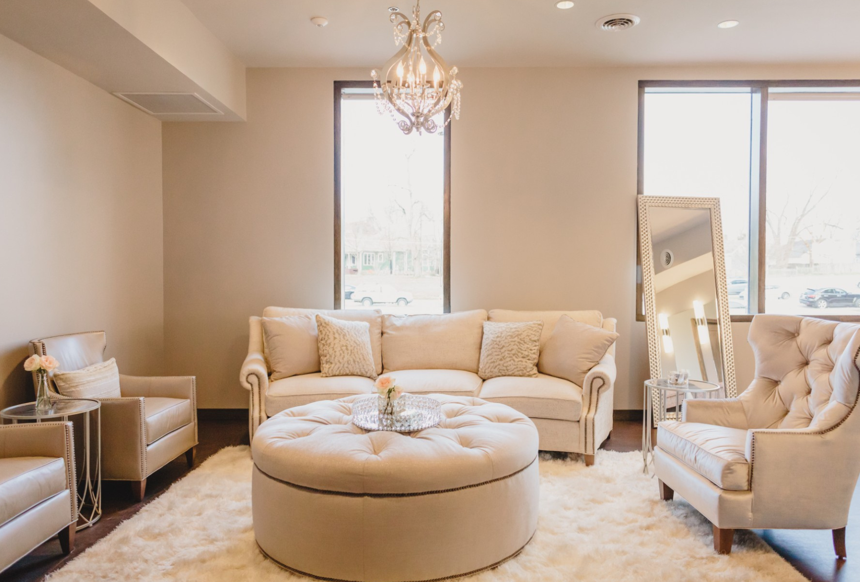 Elegant living room with cream-colored sofa, armchairs, round tufted ottoman, large mirror, chandelier, and cozy decor.