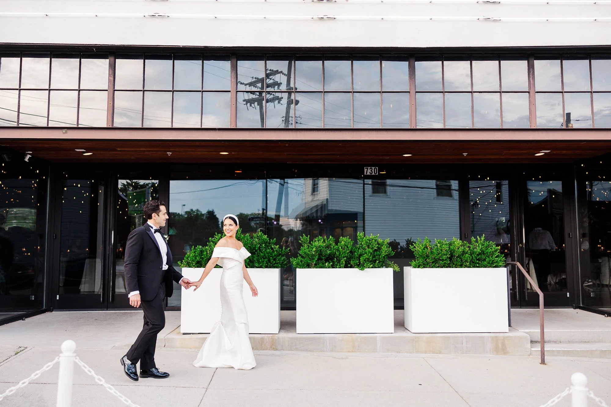 A bride and groom holding hands, walking outside a modern building with large glass windows and potted plants.