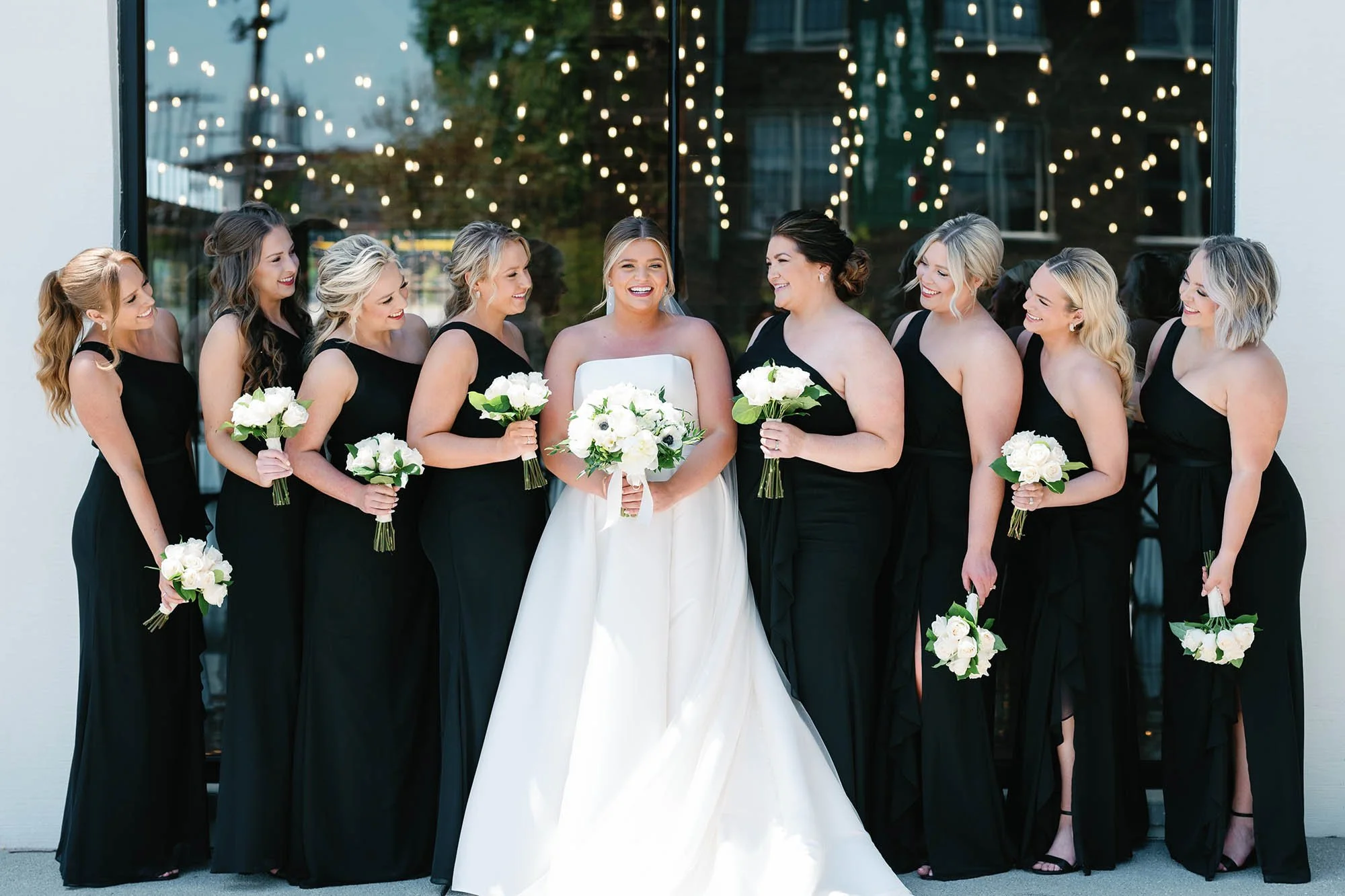 Bride in a white gown with her bridesmaids in black dresses holding white and green bouquets, standing in front of a glass window with string lights reflected in it, outdoors.
