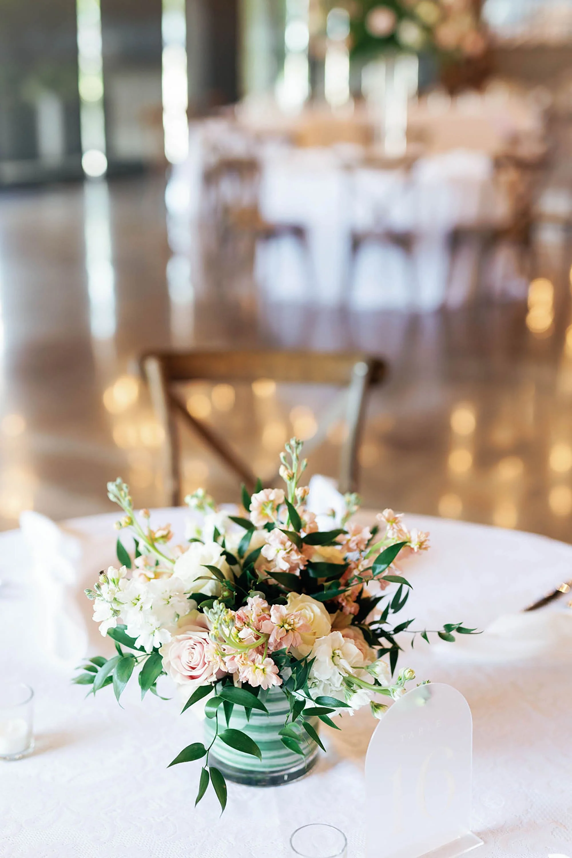 Elegant floral centerpiece with light pink roses, white flowers, and greenery on a white tablecloth at an event, with blurred background of a banquet hall and round tables.
