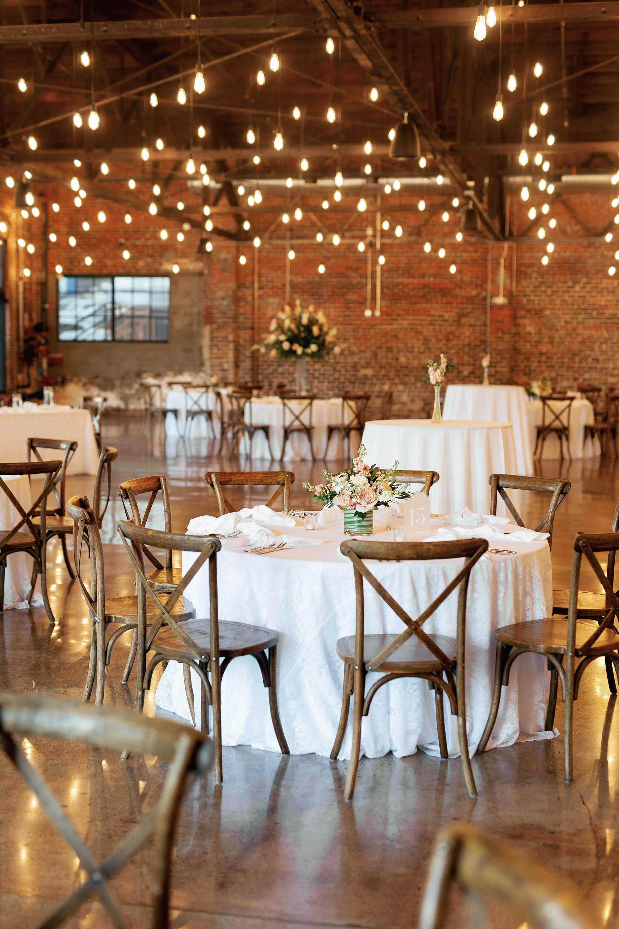 Wedding reception setup with round tables covered in white tablecloths, decorated with floral centerpieces, set with plates and cutlery. Rustic wooden chairs surround the tables. String lights hang from the ceiling in an industrial-style venue with b
