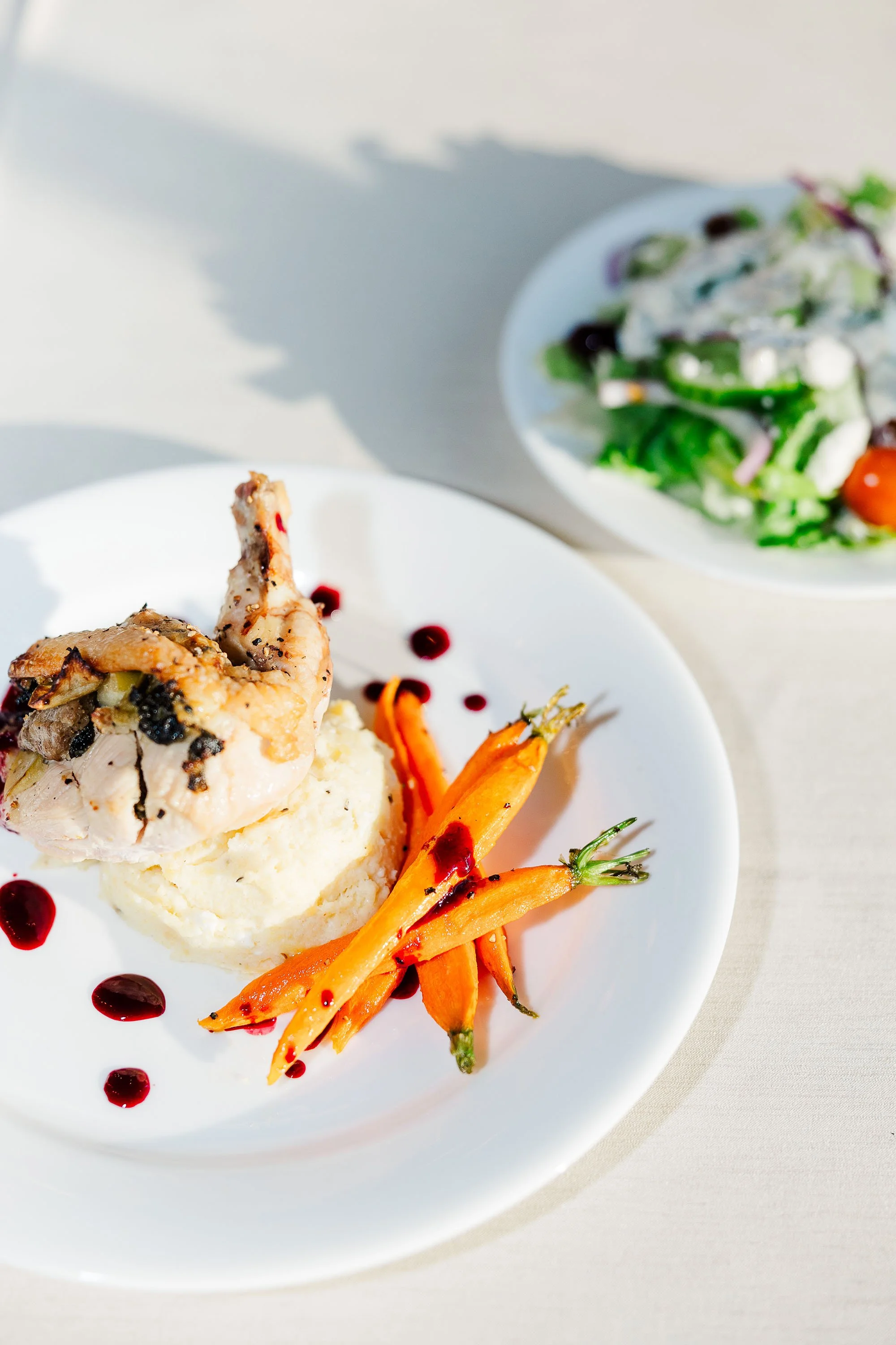 Plate with roasted chicken, mashed potatoes, and roasted carrots, drizzled with red sauce, with a side salad in the background.