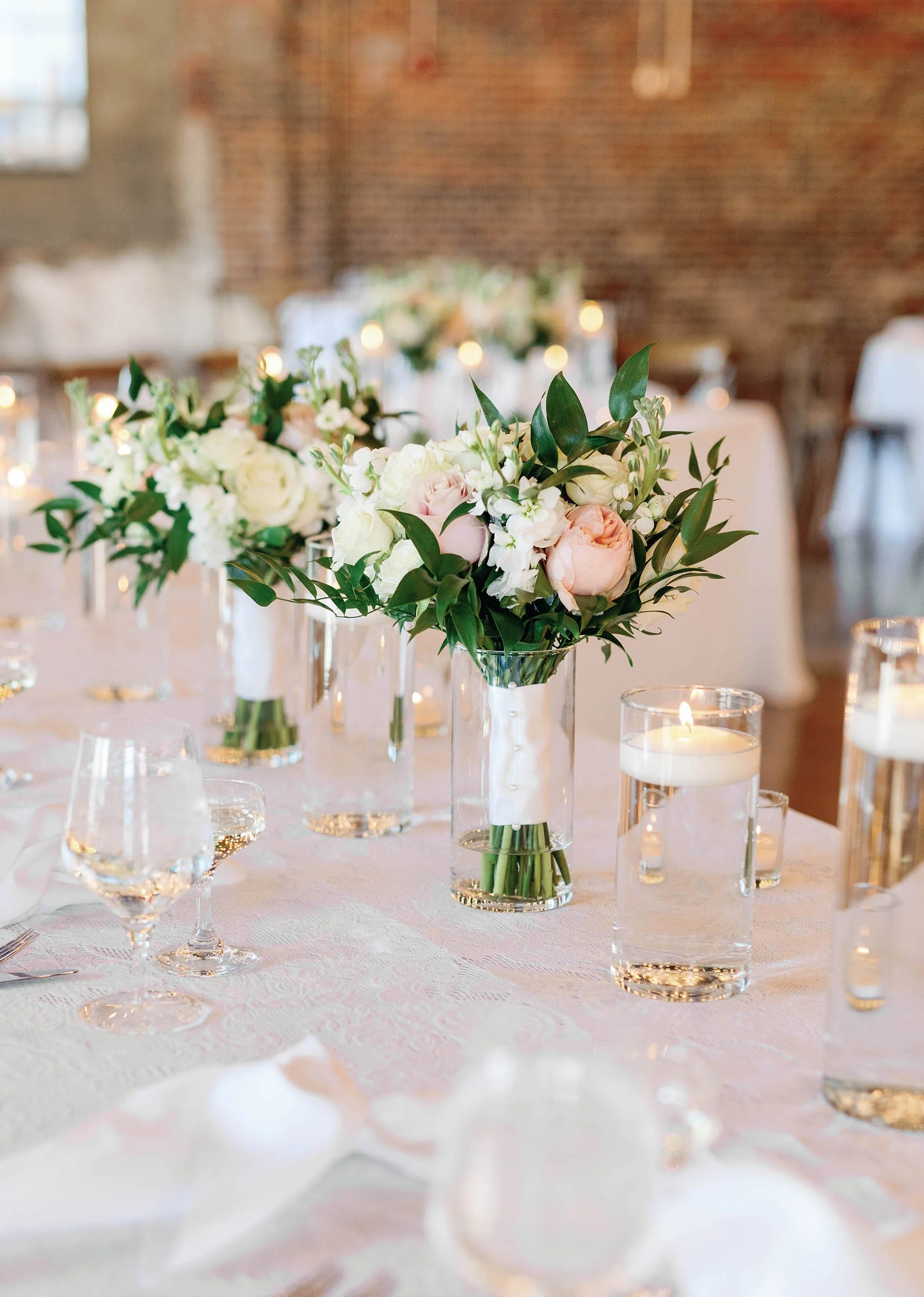 Elegant floral centerpiece with white and blush pink roses, white stock flowers, and greenery on a table with glass candle holders and candles, set in a rustic-style event space.