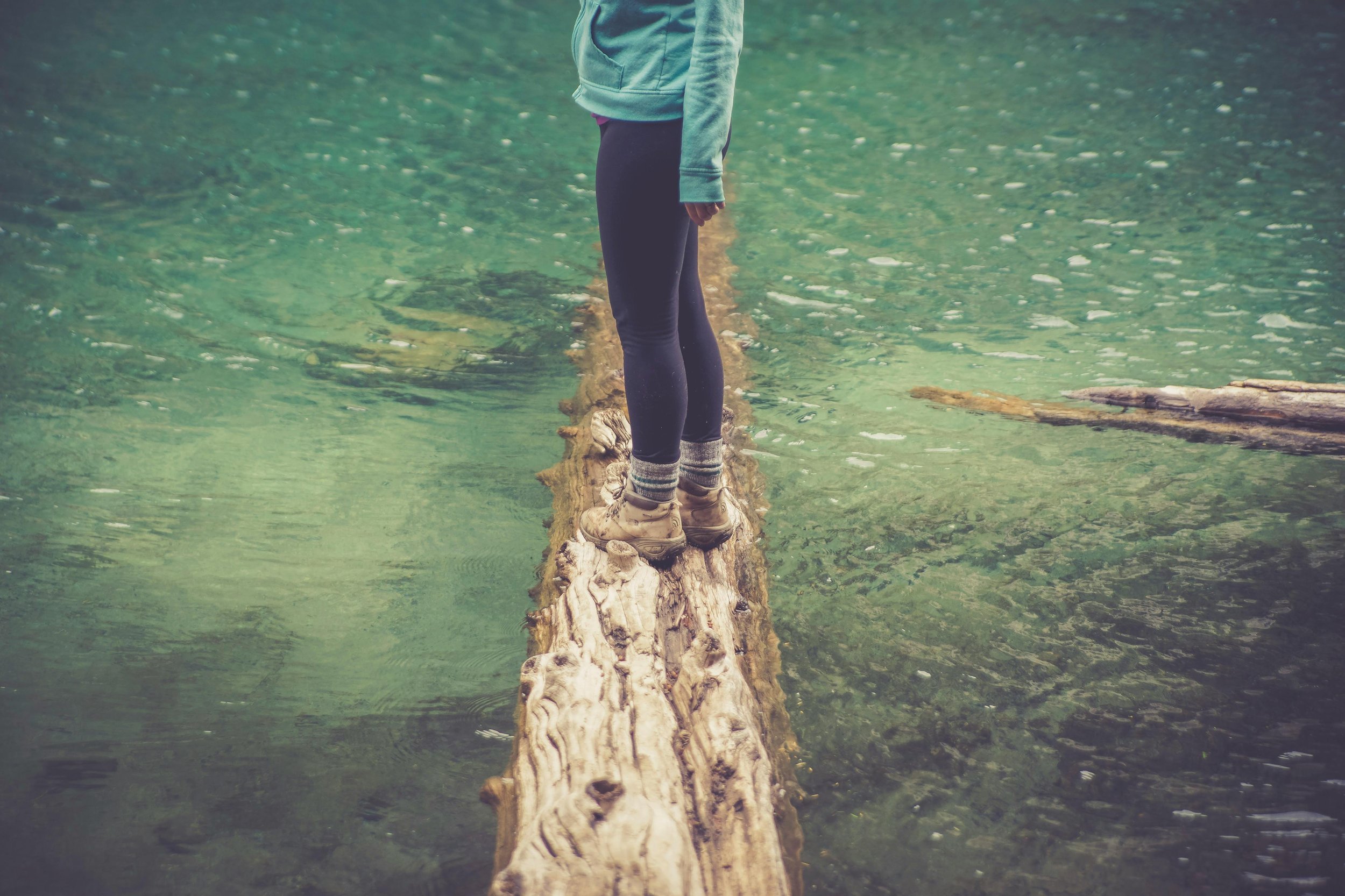 Person standing on a fallen log in a body of clear water, wearing hiking boots, leggings, and a jacket.