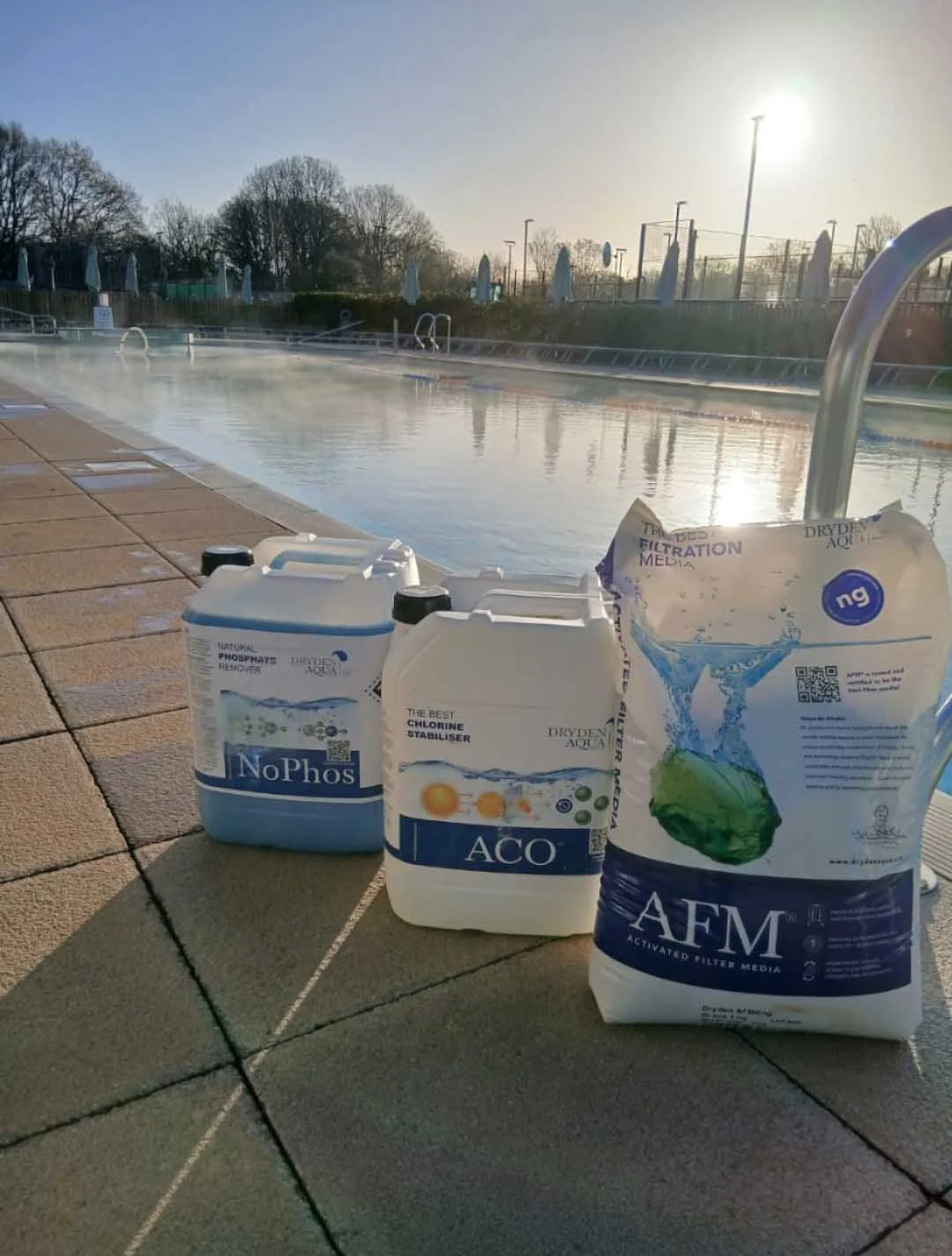 Poolside with bottles of water treatment chemicals, including NOPhos, ACO, and AFM, near a swimming pool in sunlight.